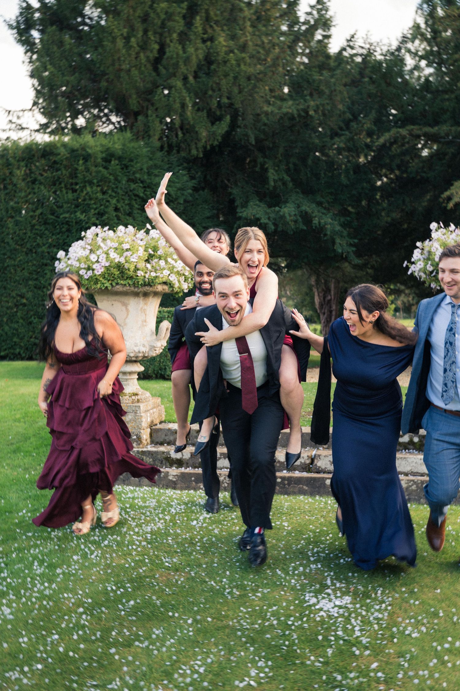 A group of wedding guests in formal attire playfully run and dance together on a garden lawn with flowers in the background.