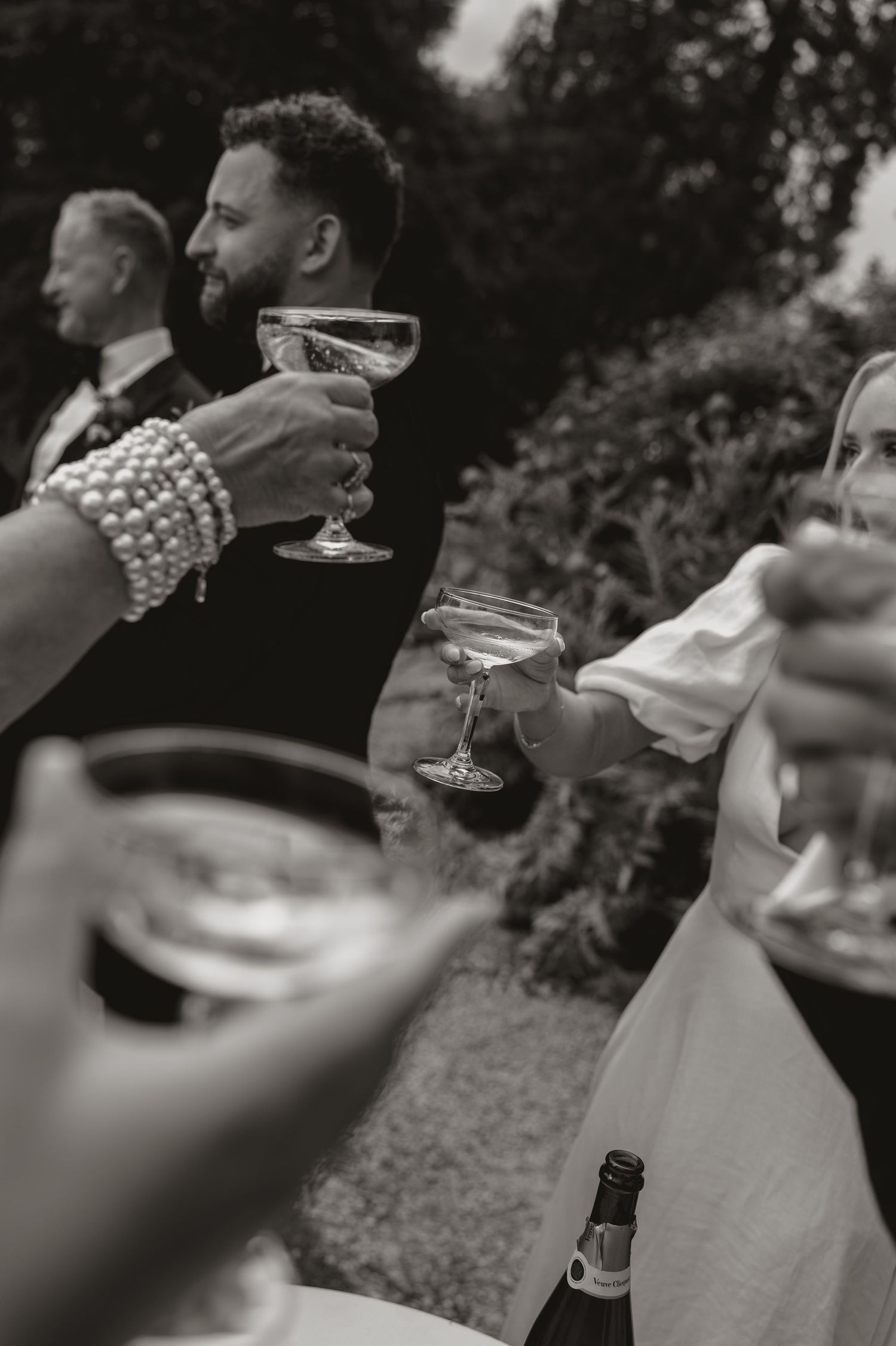 Black and white photo of champagne glasses raised in a celebratory toast at an outdoor gathering.
