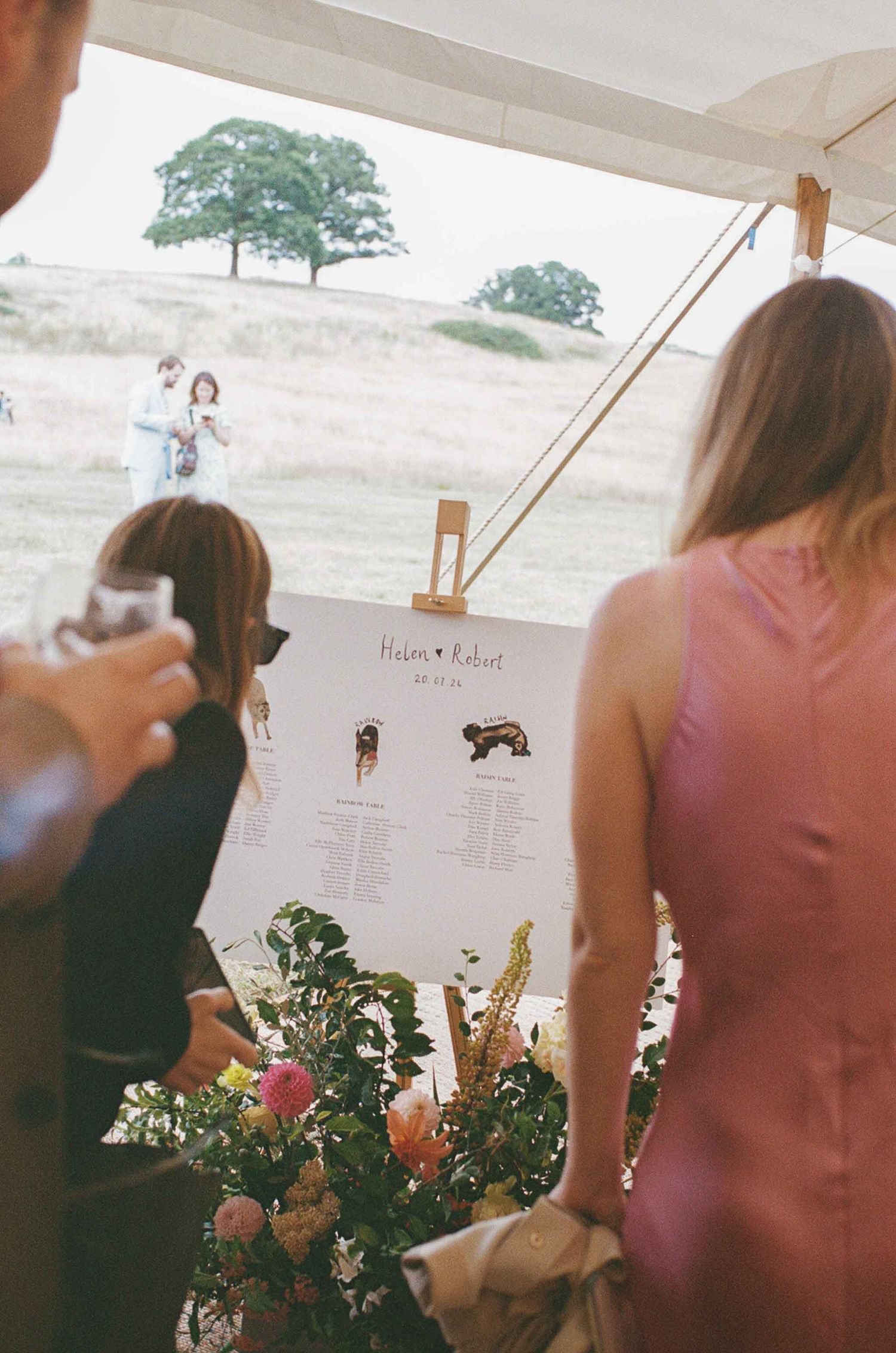 Wedding guests look through program booklets decorated with floral arrangements at an outdoor reception.