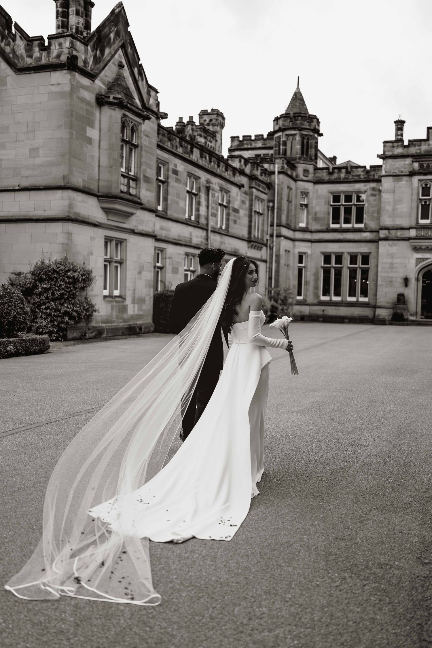 A flowing white wedding dress and long veil create dramatic movement against the backdrop of a grand gothic castle exterior.