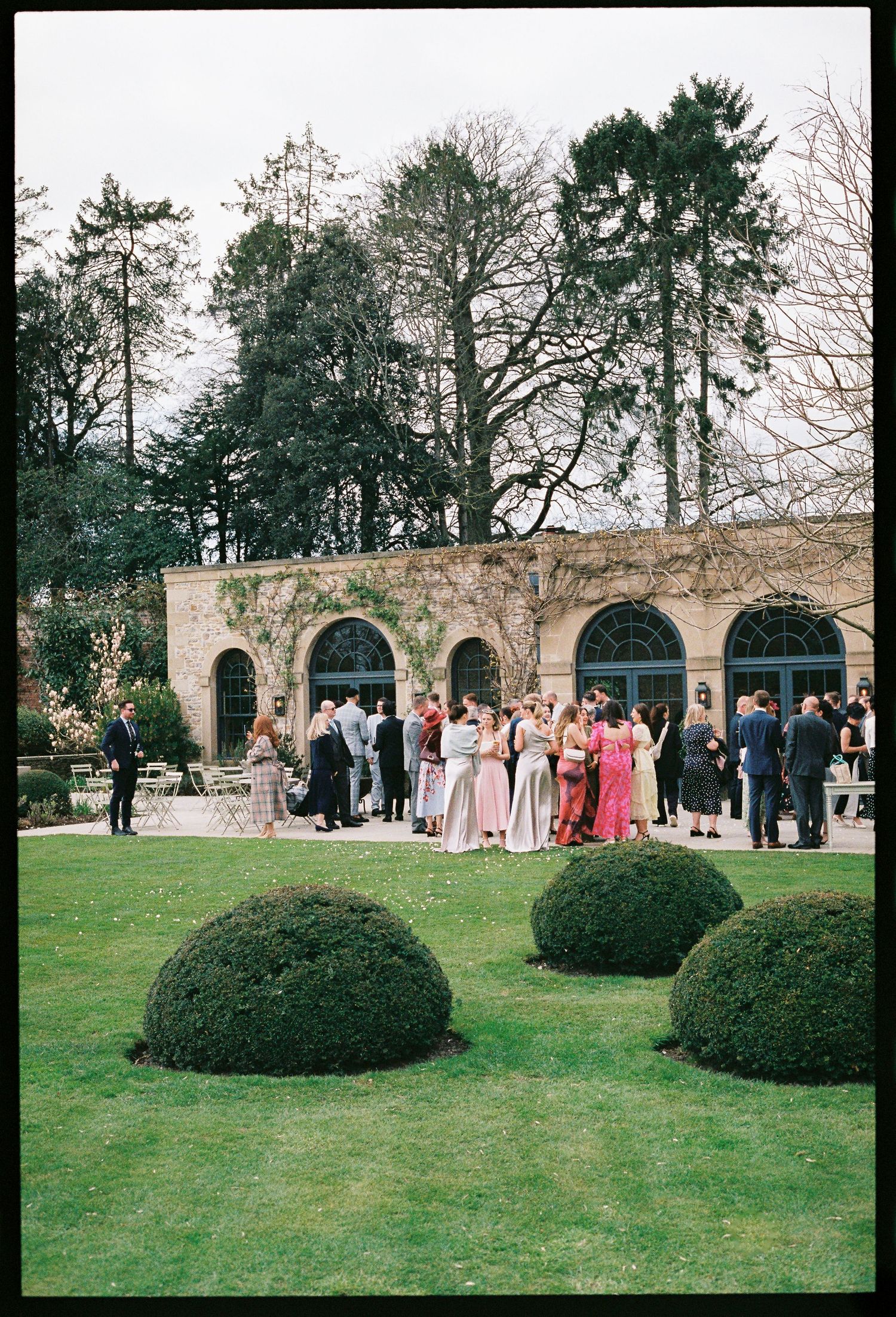Elegant outdoor wedding reception on manicured lawn with guests in formal attire outside a stone venue with arched windows.