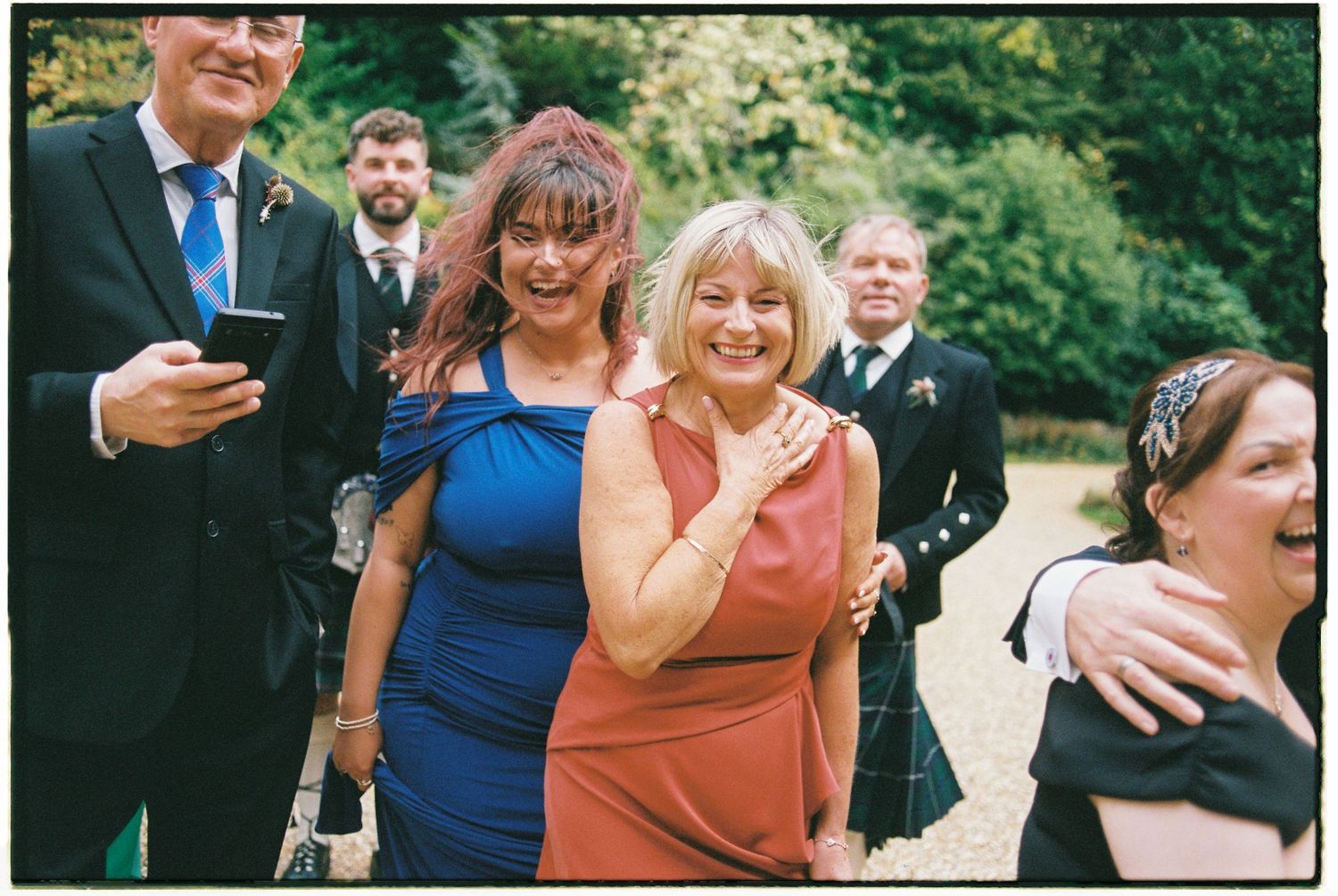A group of wedding guests in formal attire smile and laugh together during an outdoor celebration.
