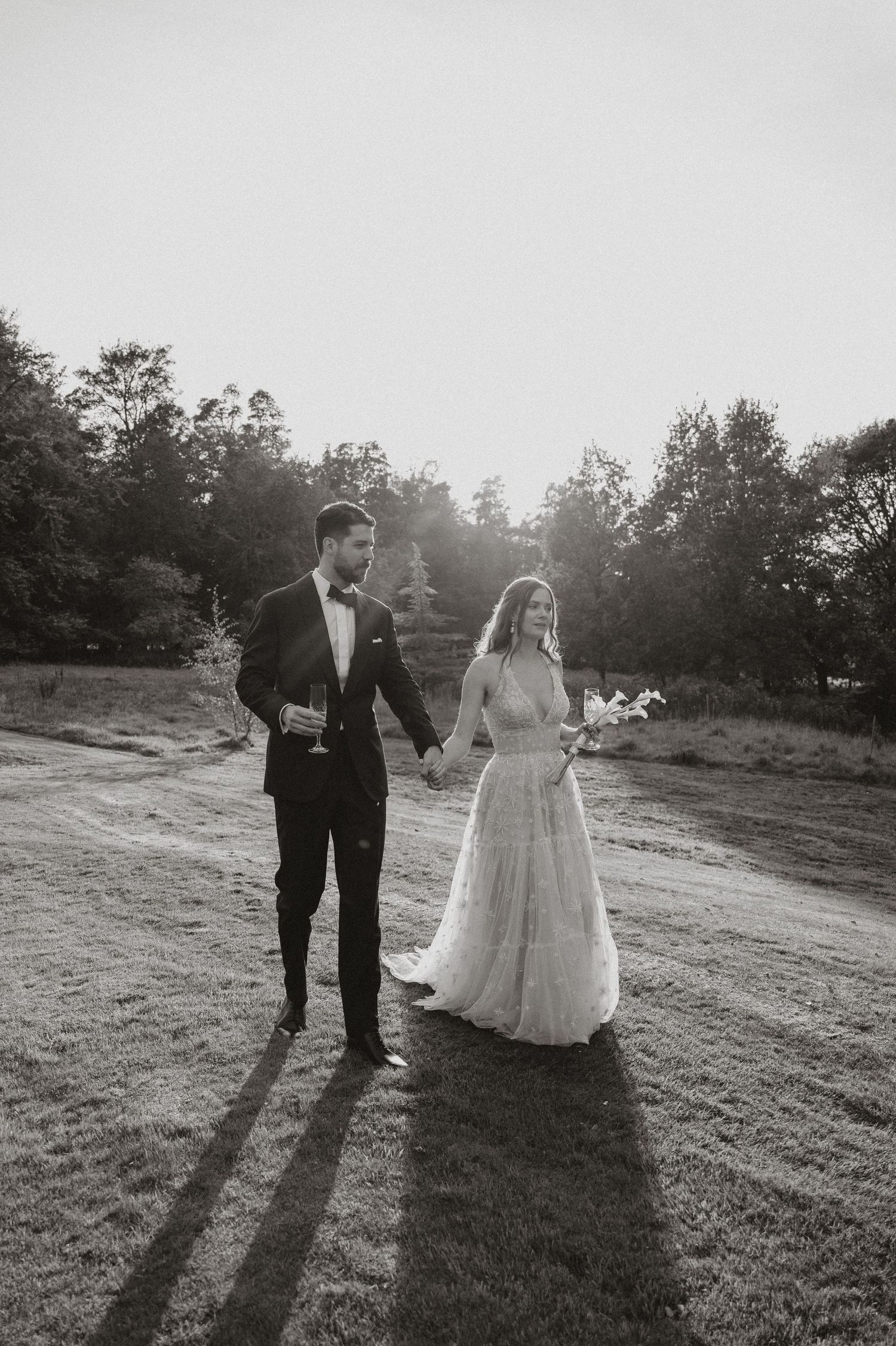 Black and white photo of a bride and groom holding hands while standing on a grassy hill at sunset.