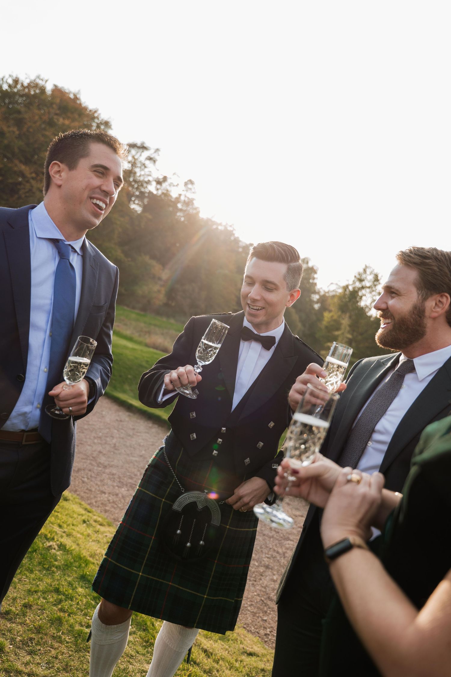 A group of groomsmen in formal attire share drinks and laughter during an outdoor wedding celebration at sunset.