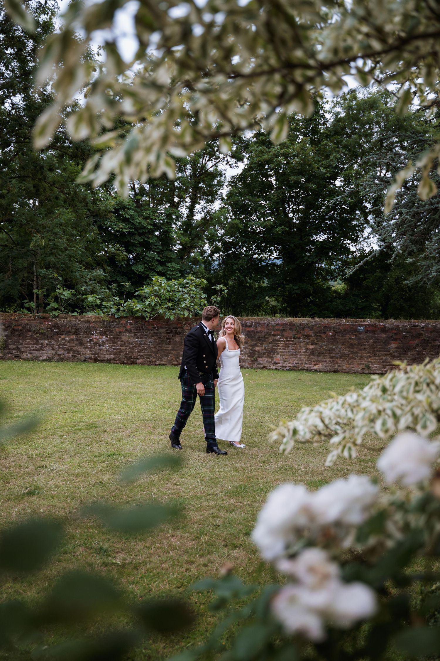 A couple in wedding attire stands together on a grassy lawn framed by delicate white blossoms in the foreground.