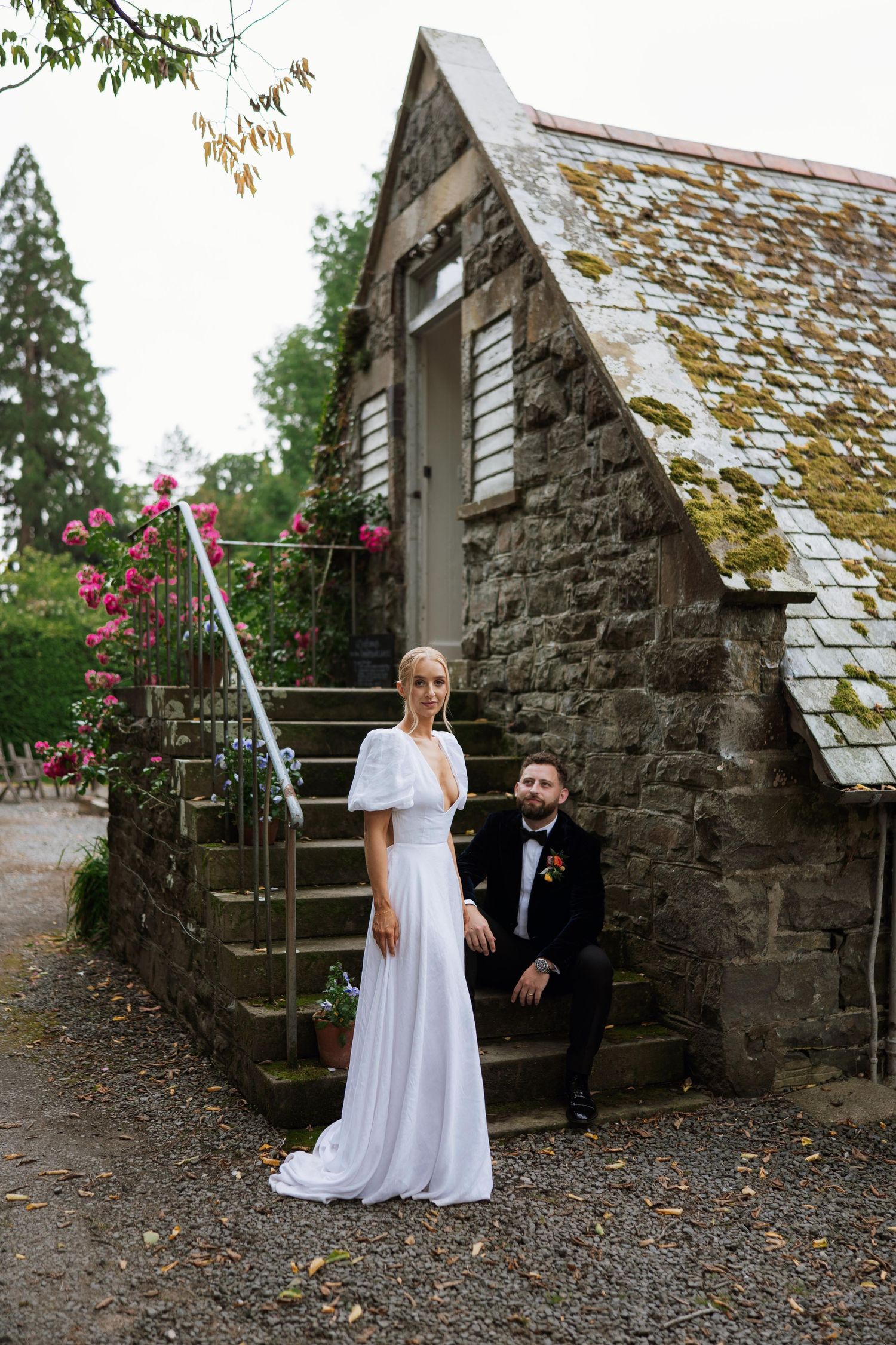A wedding couple poses outside a rustic stone cottage with a moss-covered roof and pink climbing roses.