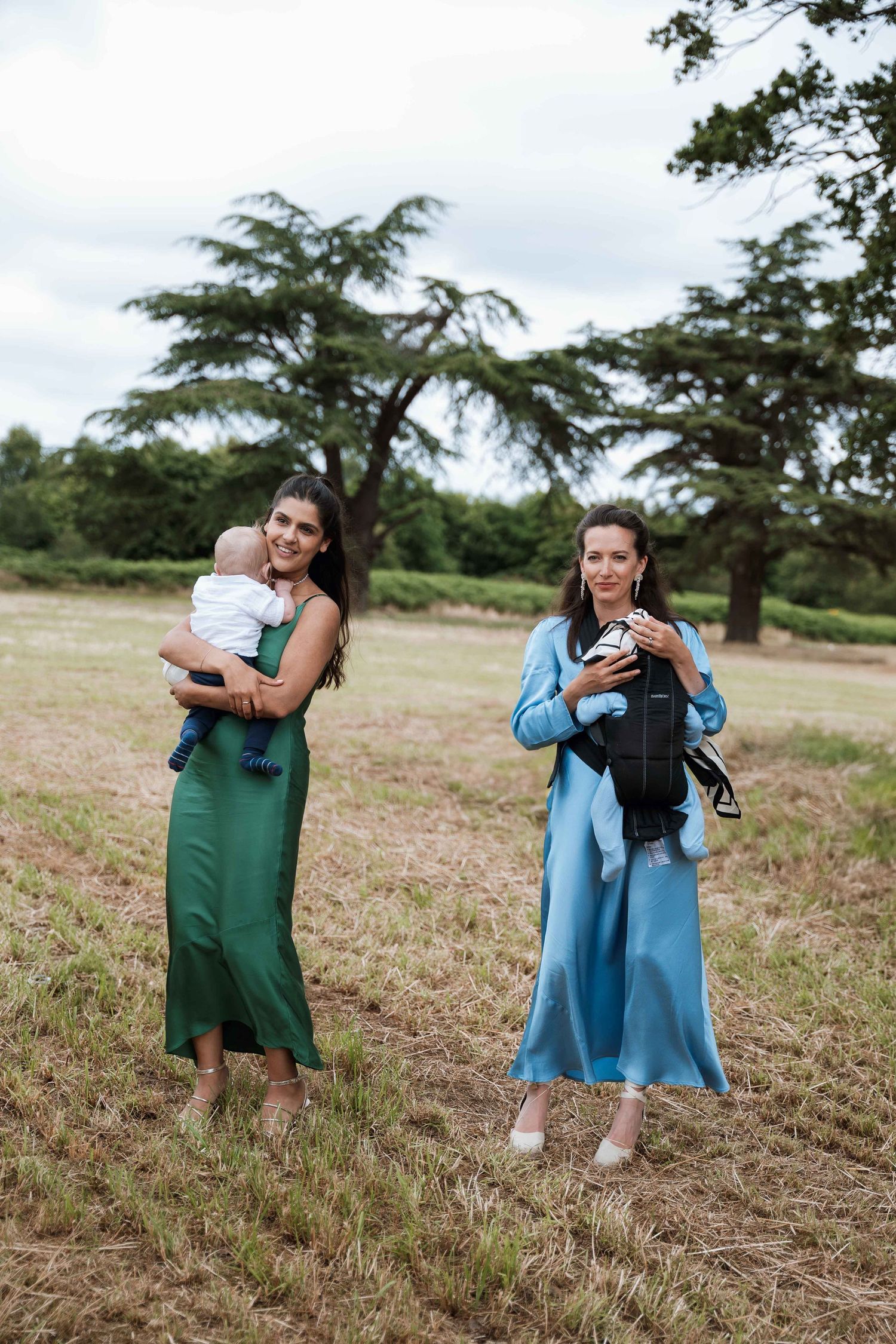Two people embrace in a grassy field on a cloudy day wearing blue and green dresses.