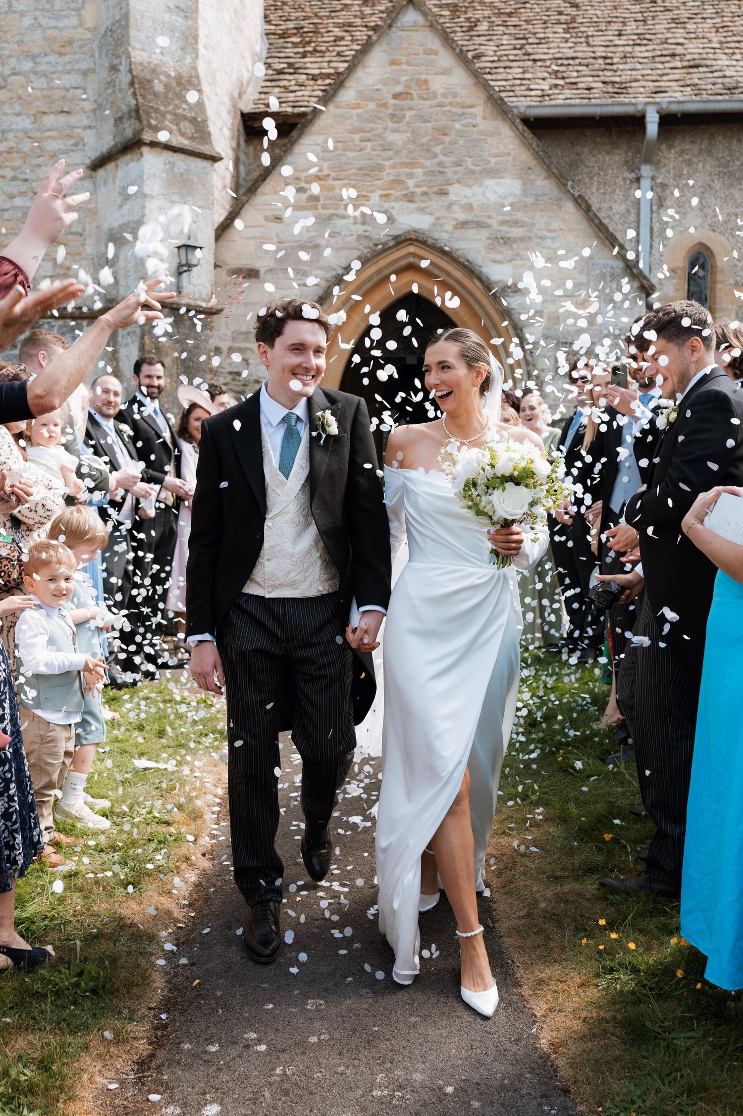 Wedding party celebrates with confetti outside a stone church as newlyweds walk through a joyful shower of white petals.