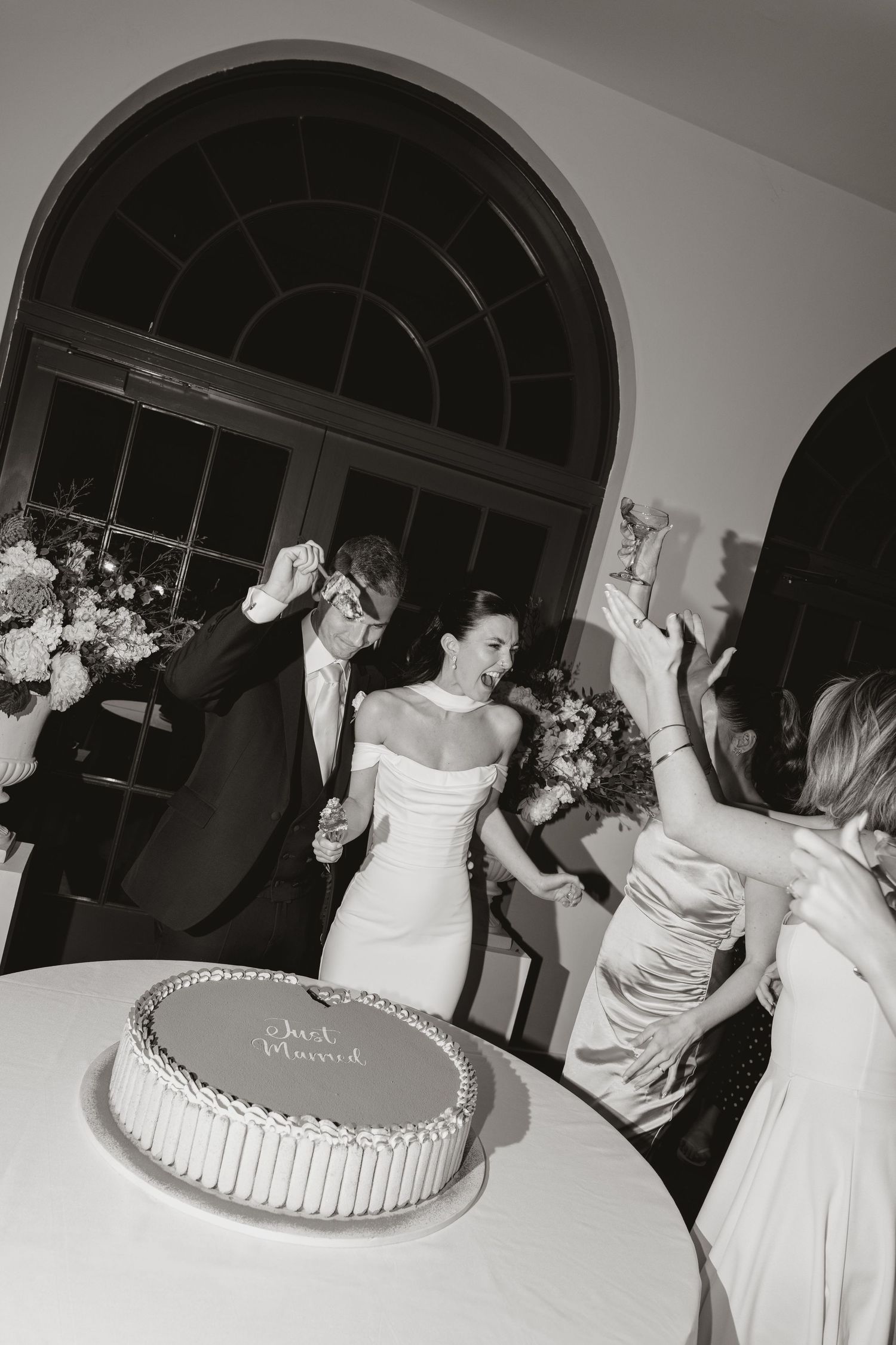 Black and white photo of newlyweds celebrating beside a Just Married wedding cake at their elegant reception.
