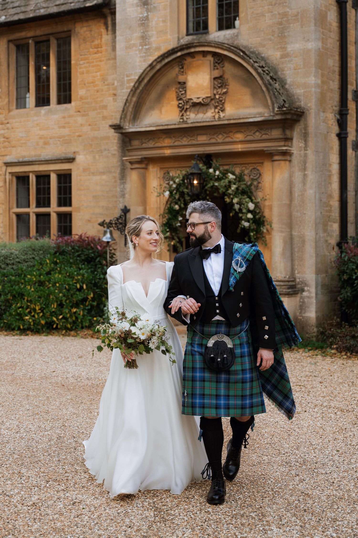 A couple walks along a gravel path outside a grand stone building with traditional Scottish wedding attire.