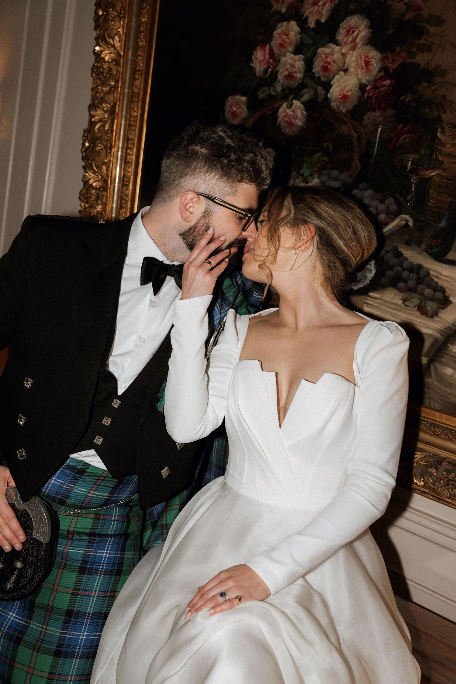 A candid wedding moment showing a couple laughing together in front of a floral painting, with the groom in a kilt.