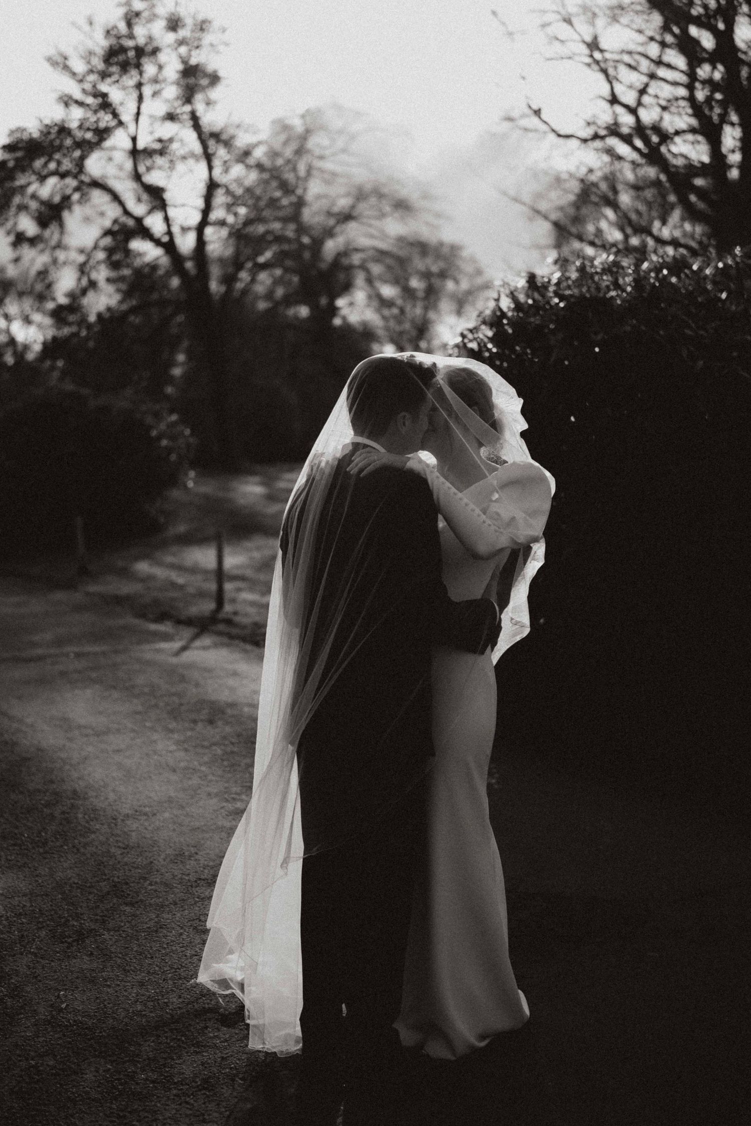 A romantic black and white wedding photograph shows a couple embracing on a garden path with flowing veil and trees silhouetted.