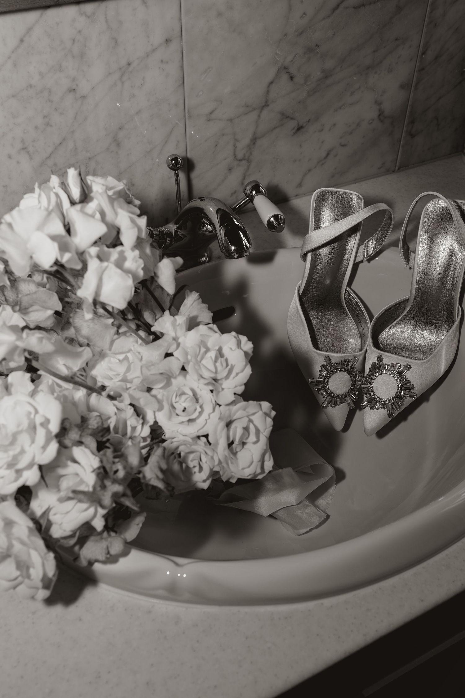 Black and white photo of elegant bridal shoes with crystal buckles next to white roses on marble surface. wedding bridal details.