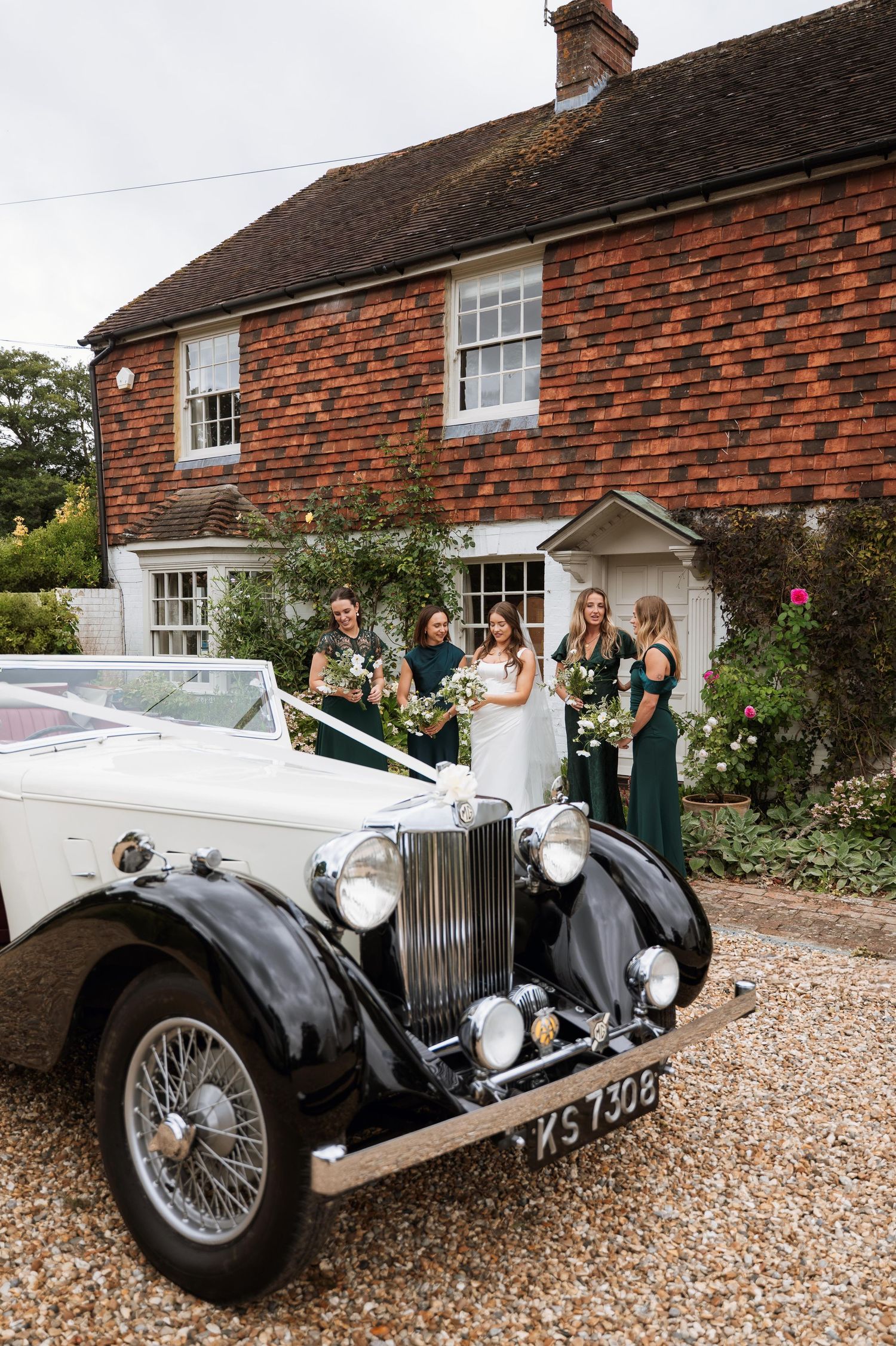 Vintage silver Rolls Royce classic car parked on gravel driveway in front of traditional English brick house.
