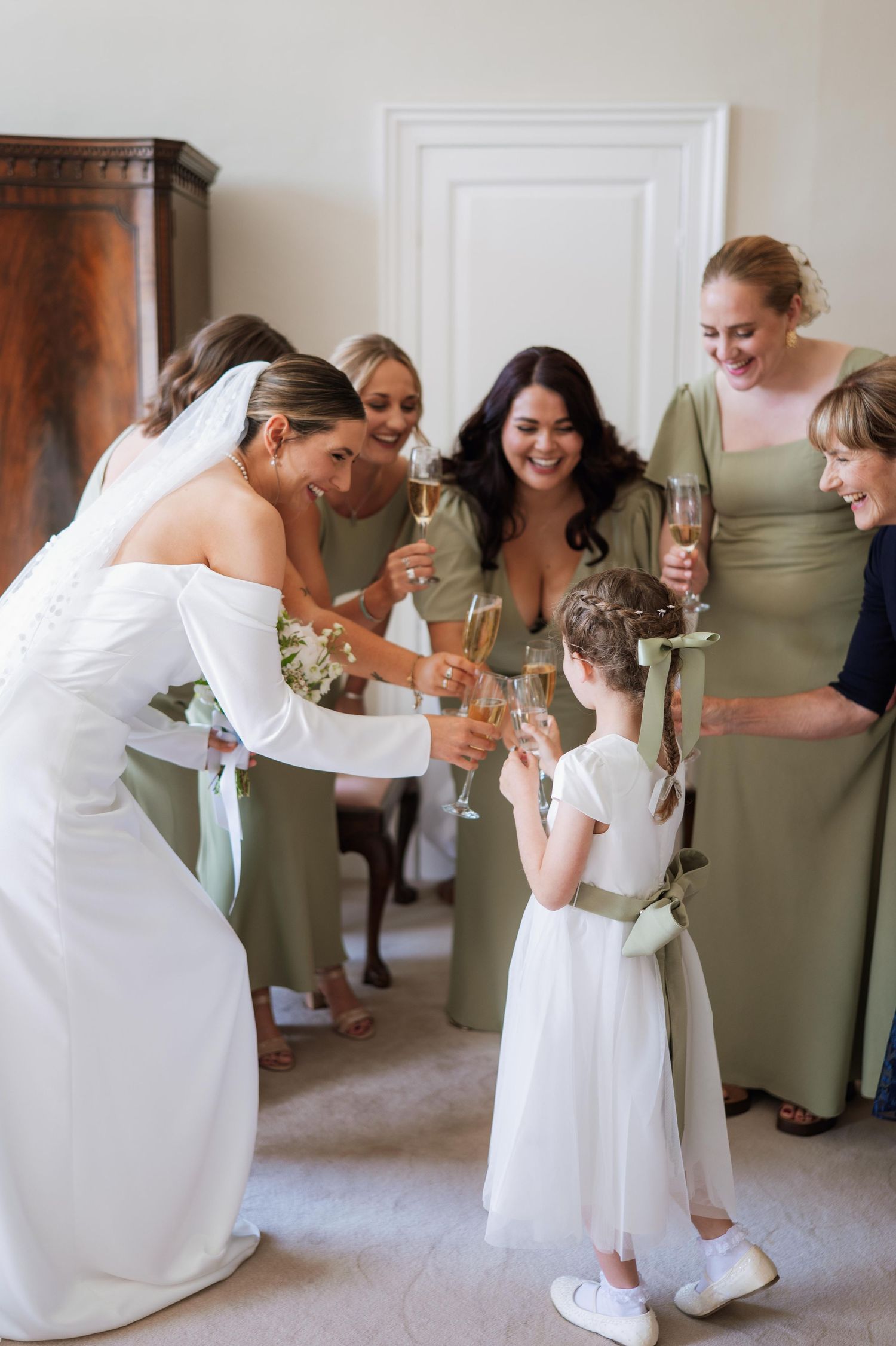 A bride in a white dress and bridesmaids in sage green dresses share joyful moments during wedding preparations.