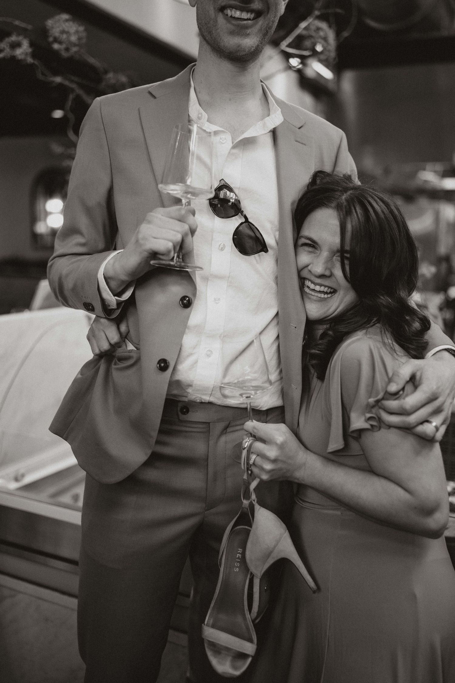 A black and white candid photo of a couple sharing a joyful moment at a party, holding drinks and shoes.