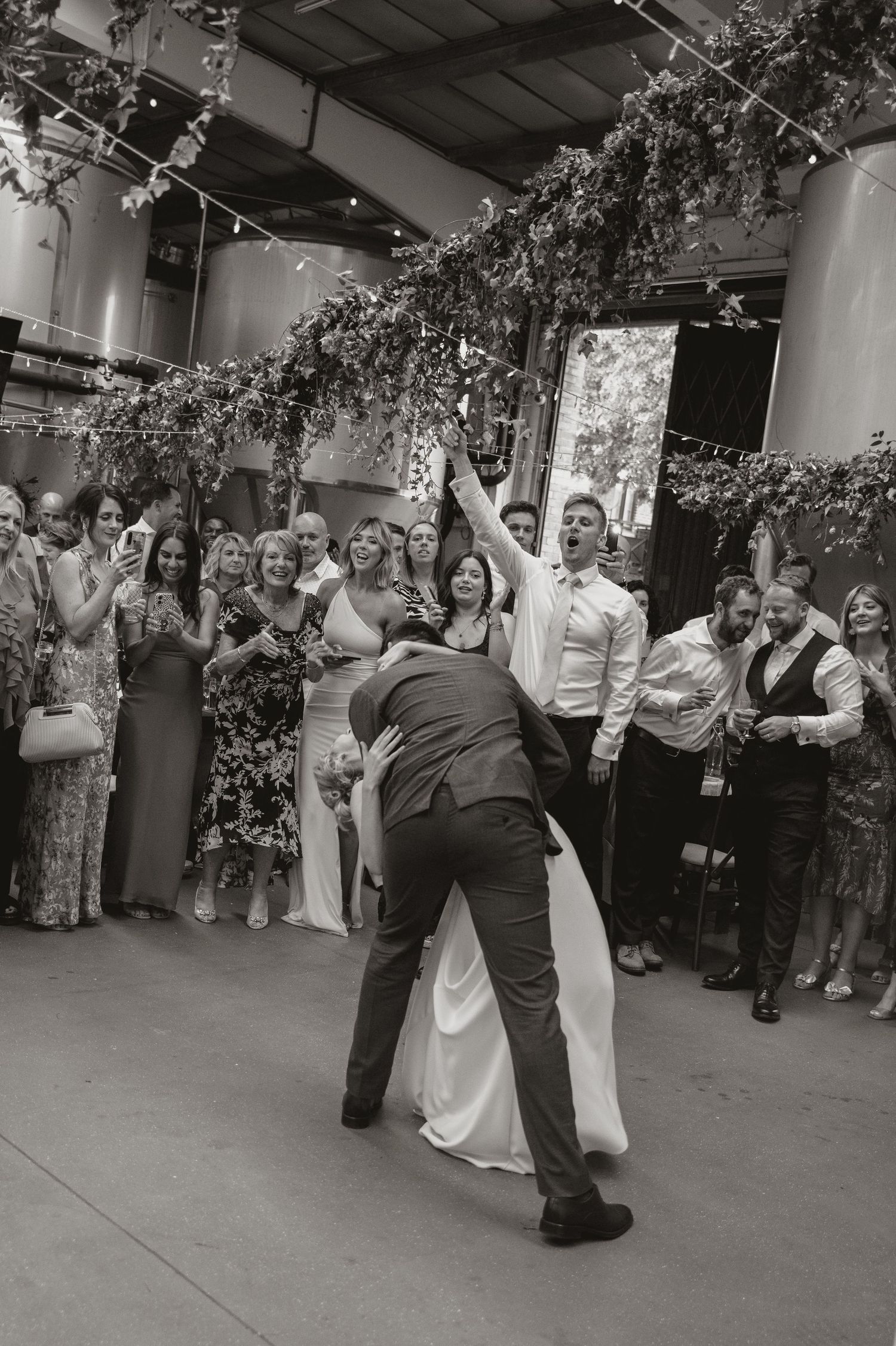 A couple performs their first dance at a wedding reception under hanging greenery and string lights while guests watch.