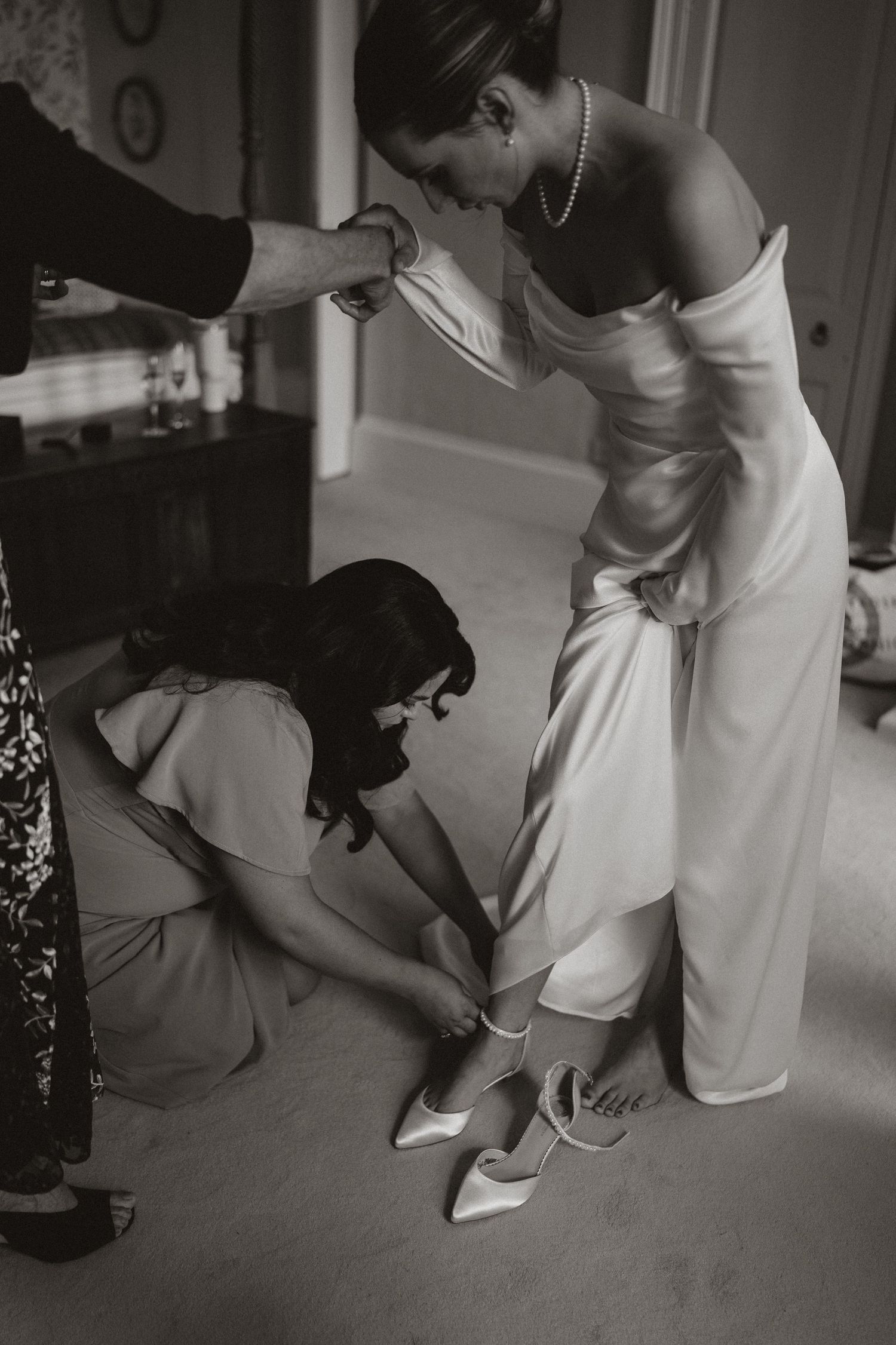 A vintage black and white photograph shows a bride getting help putting on her white wedding shoes while wearing an elegant gown.