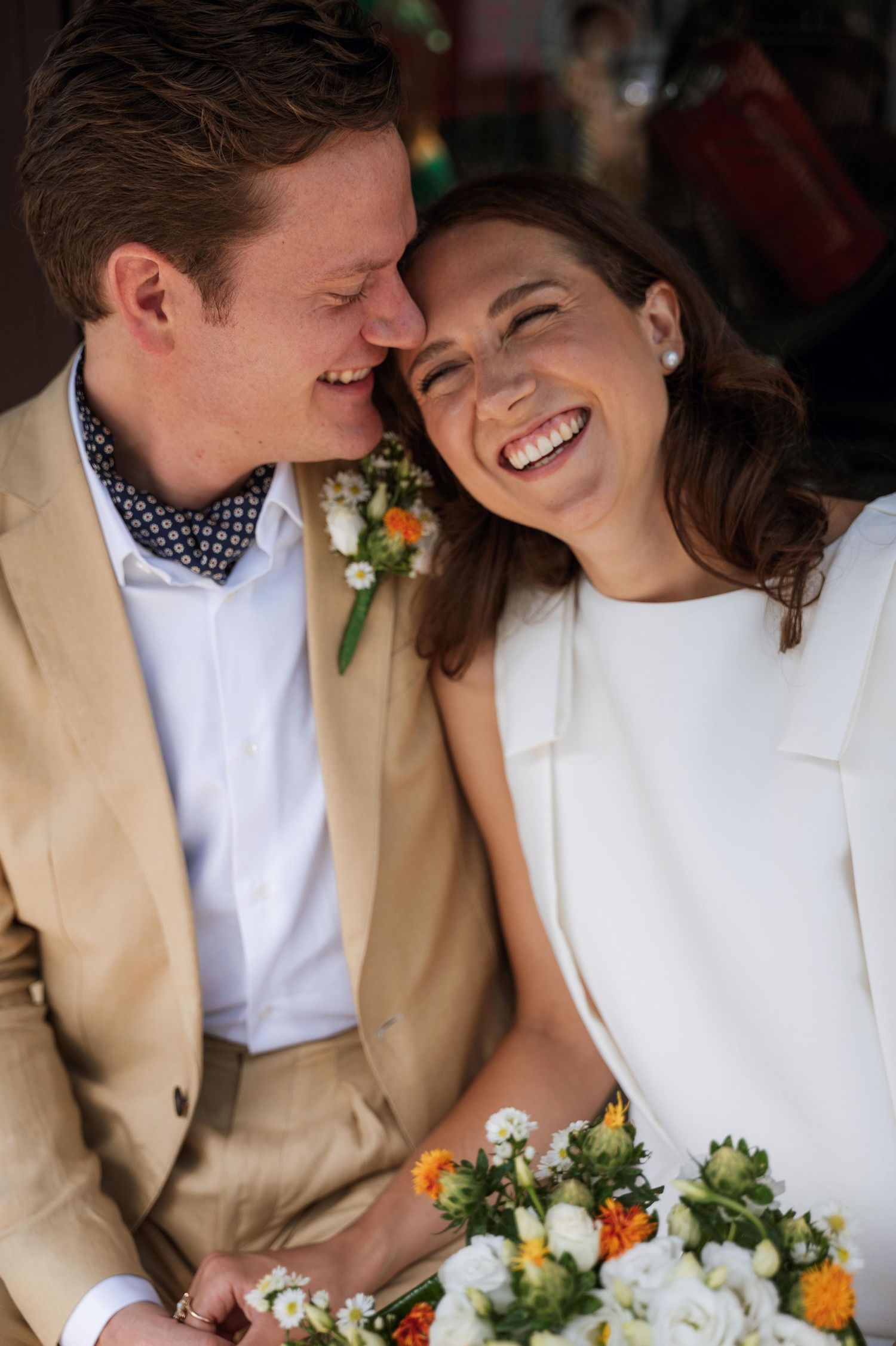 A couple shares joyful laughter while sitting together in formal wedding attire with a bouquet of white and orange flowers.