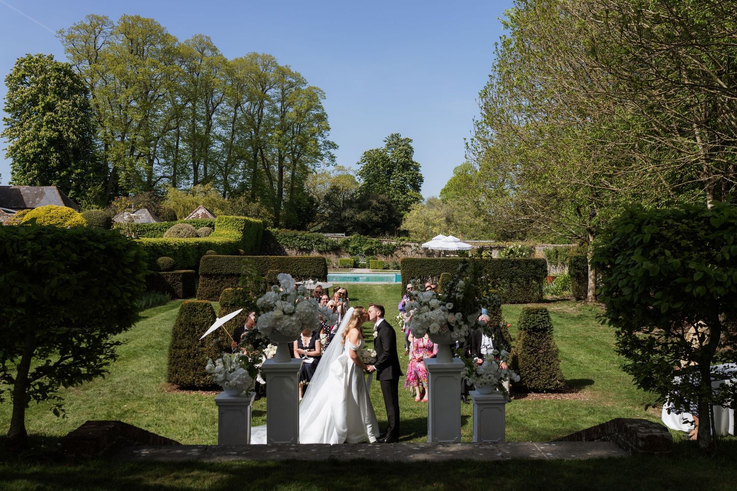 Bride and groom walk through elegant formal garden with sculpted hedges and statues on a sunny spring wedding day.