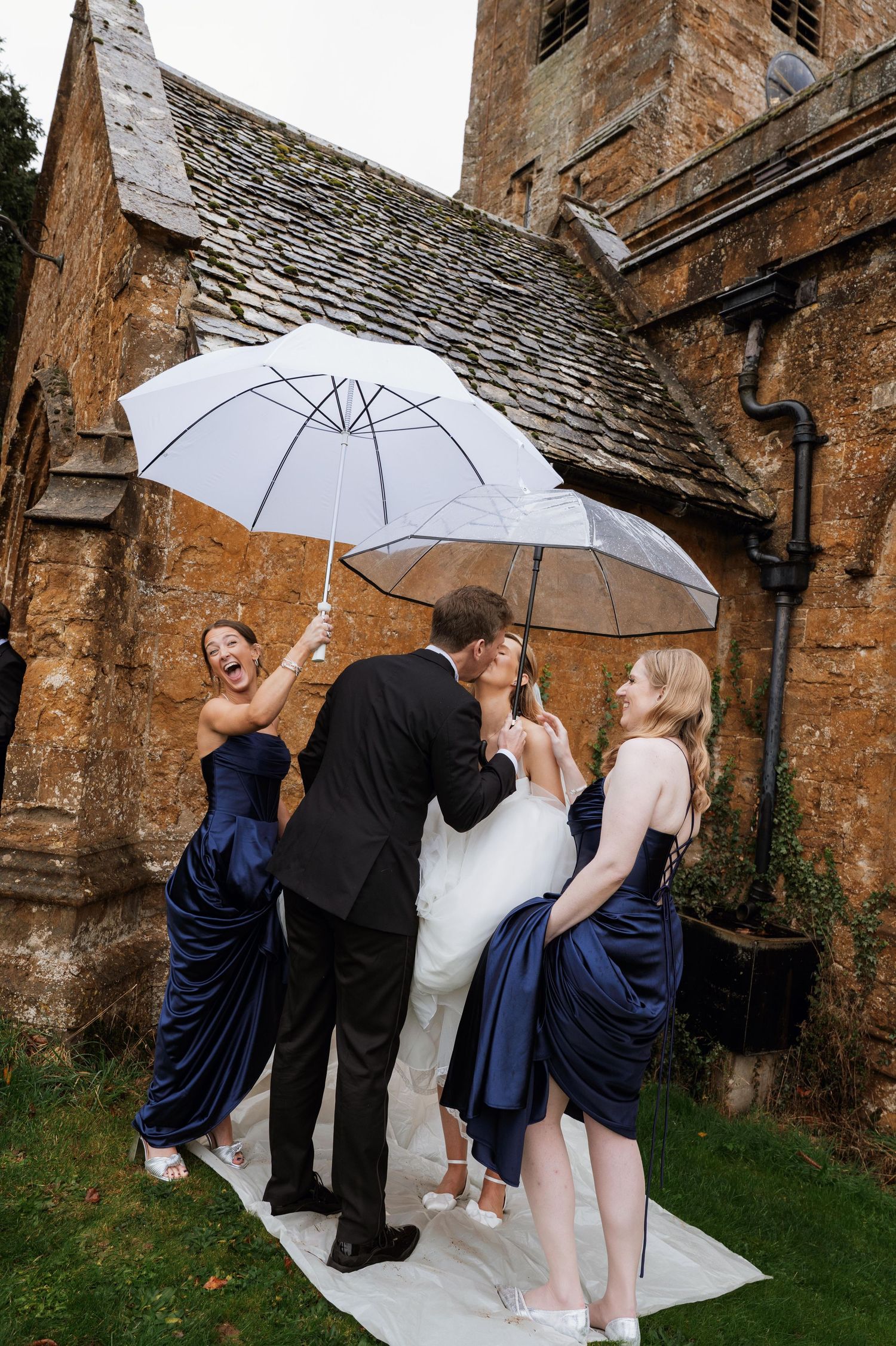 A wedding party stands under white umbrellas outside a rustic stone church with a weathered roof.