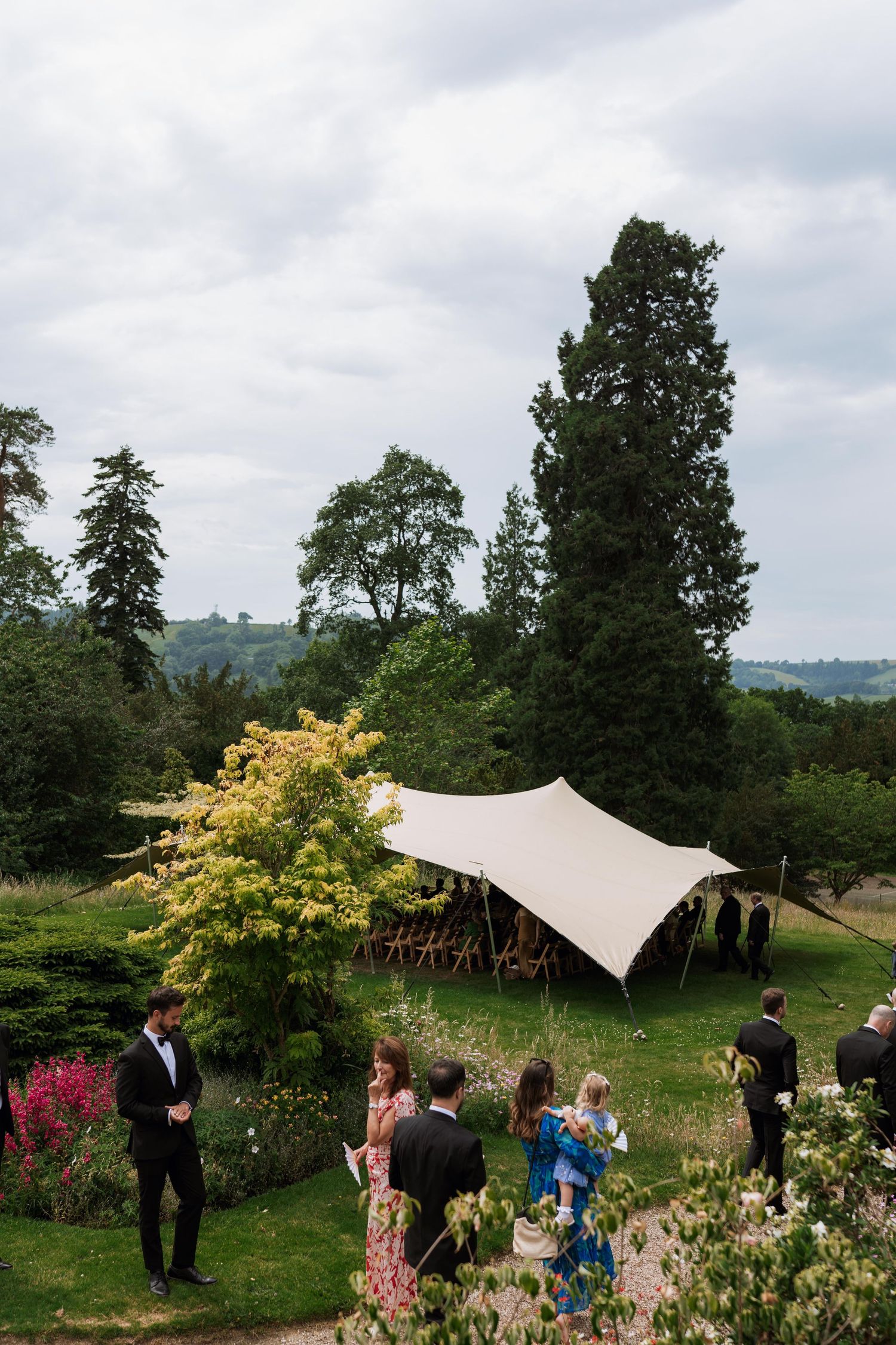 Wedding guests gather under an elegant white canopy tent in a lush garden setting with blooming flowers and trees.