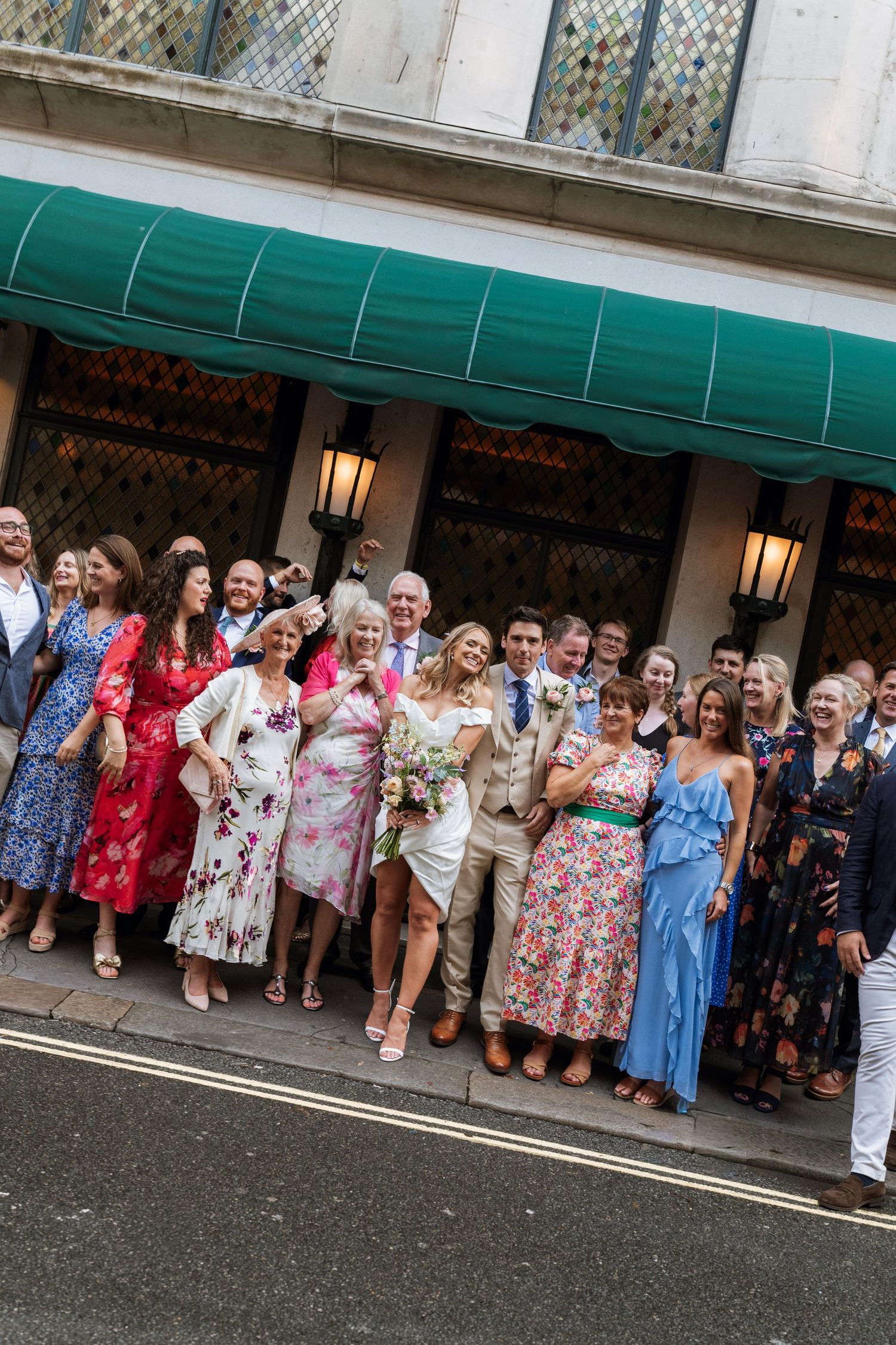 A large group of wedding guests in colorful floral dresses and formal attire gather under a green awning outside a building.