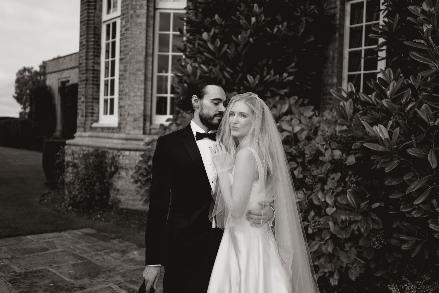 Two people embrace and share a loving moment in front of a rustic stone building with lush greenery surrounding them.