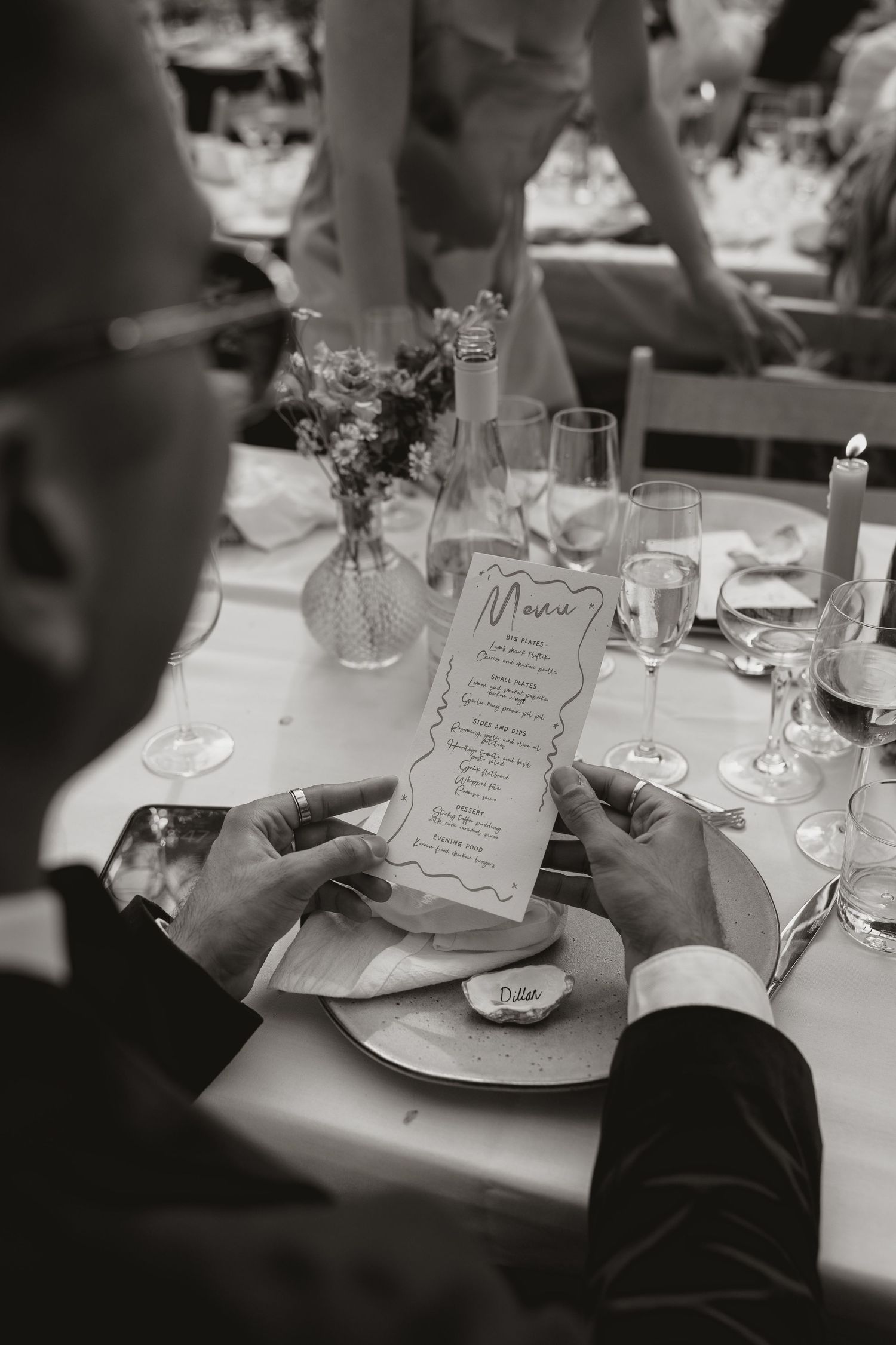 Black and white photo of hands holding an elegant menu card at a formal dinner table with wine glasses and floral decor.