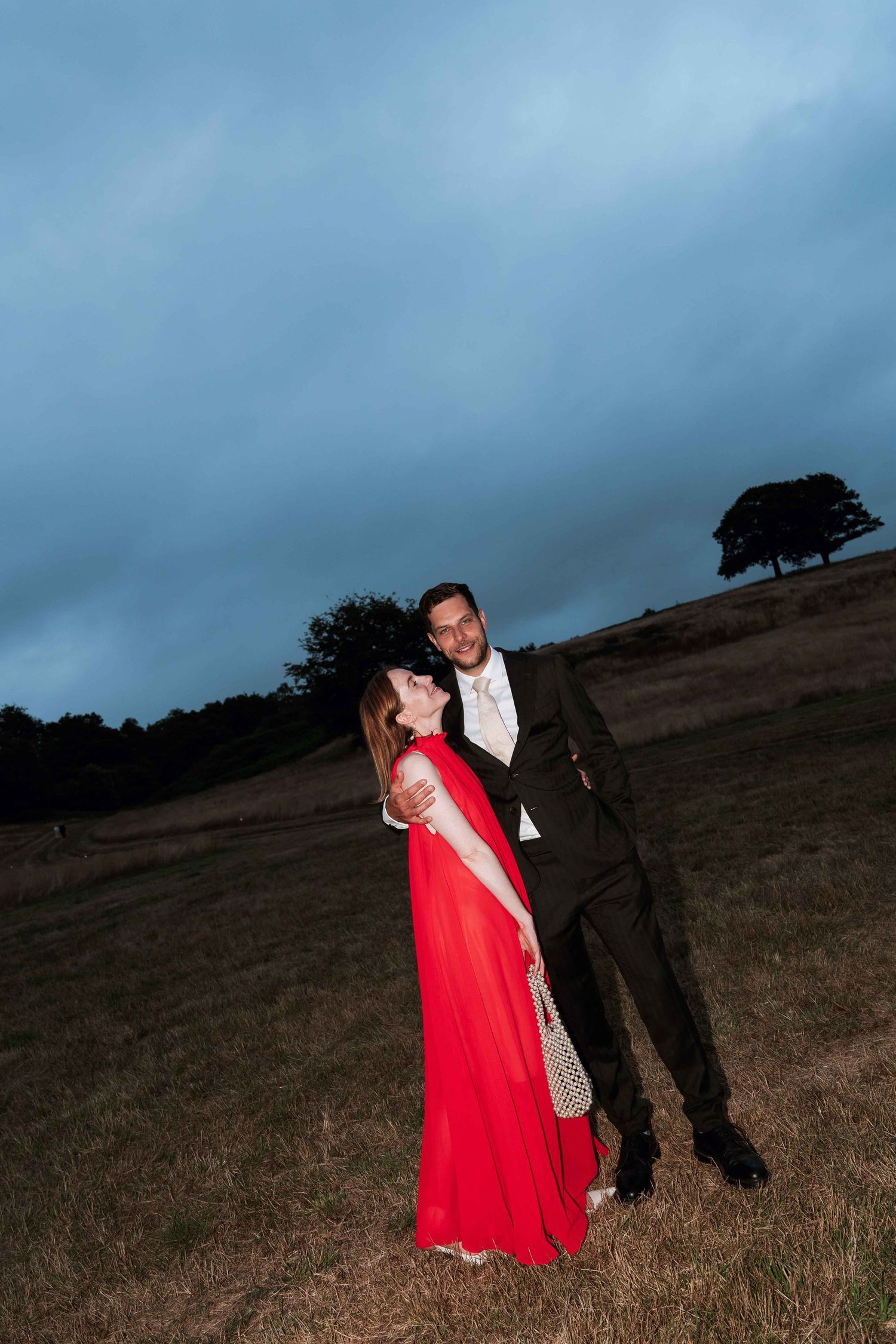 A couple in evening attire poses dramatically against a moody hillside landscape at dusk with a lone tree silhouette.