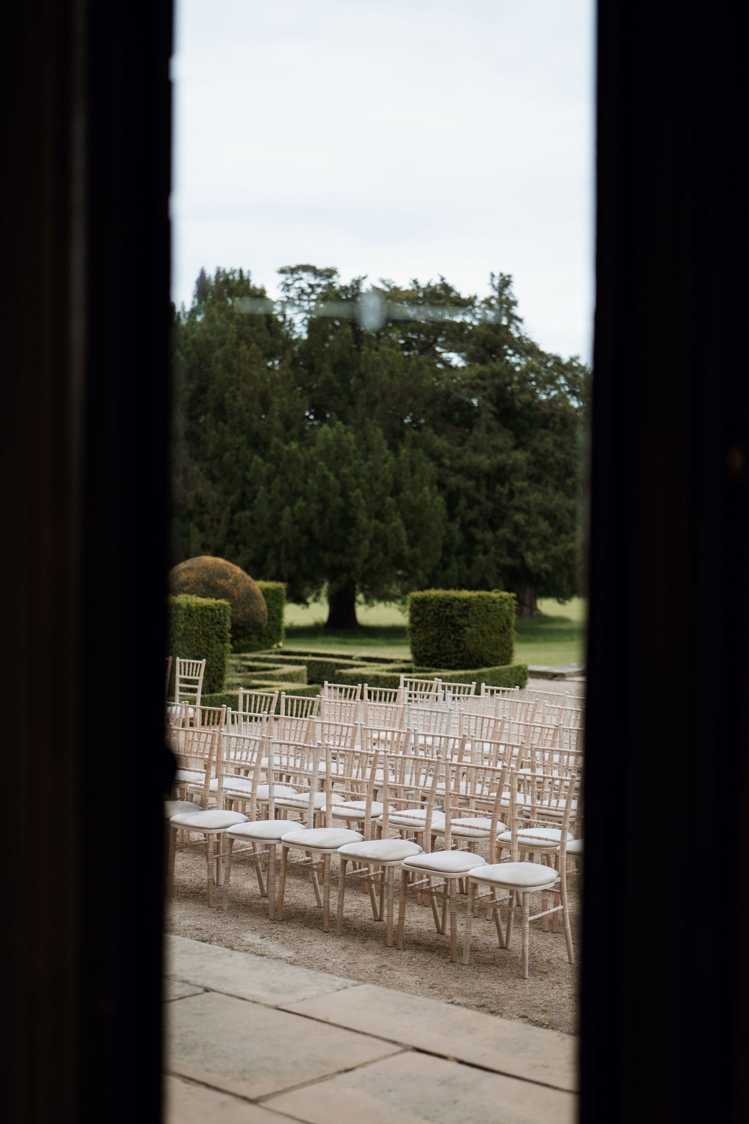 White wooden chairs arranged in rows for an outdoor wedding ceremony on a stone patio with trees in the background.