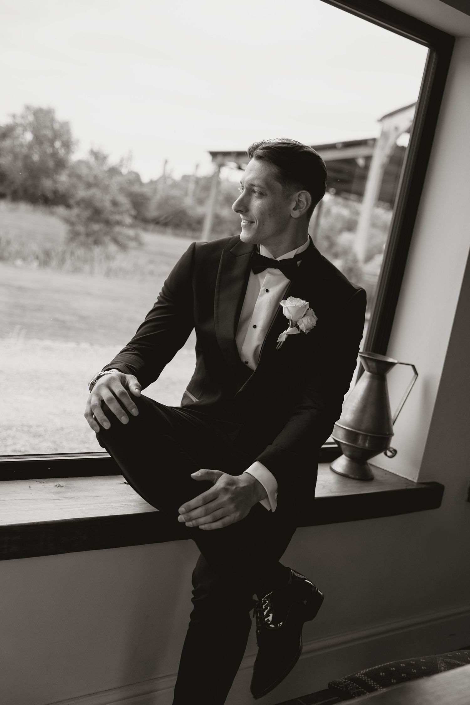 Groom in black tuxedo with boutonniere sits on windowsill, smiling, black and white wedding photo.