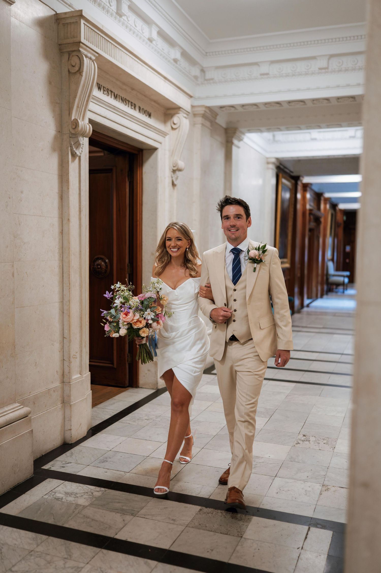 Wedding couple playfully runs through an elegant marble hallway with white columns and wooden doors.