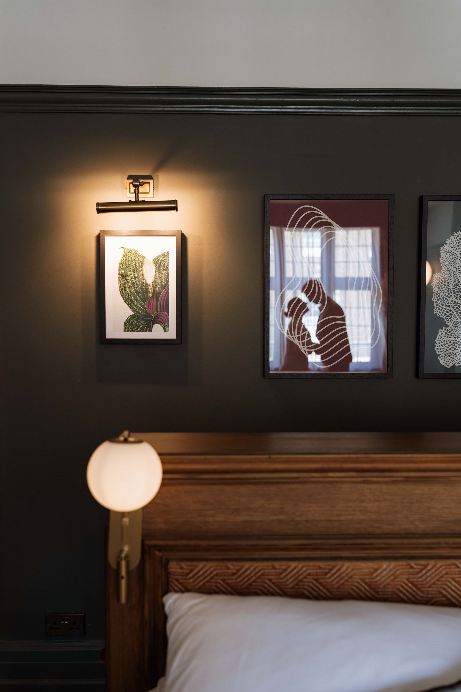A dark wooden bedroom wall features illuminated artwork and a lamp casting warm light over a rustic headboard.