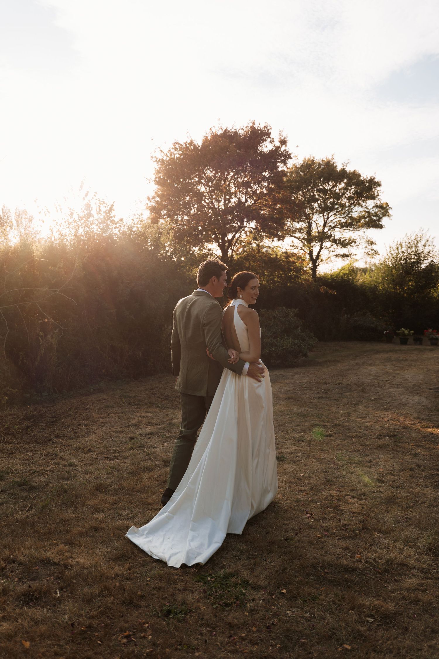 A couple in wedding attire walks along a hillside at golden hour as sunlight streams through trees in the background.