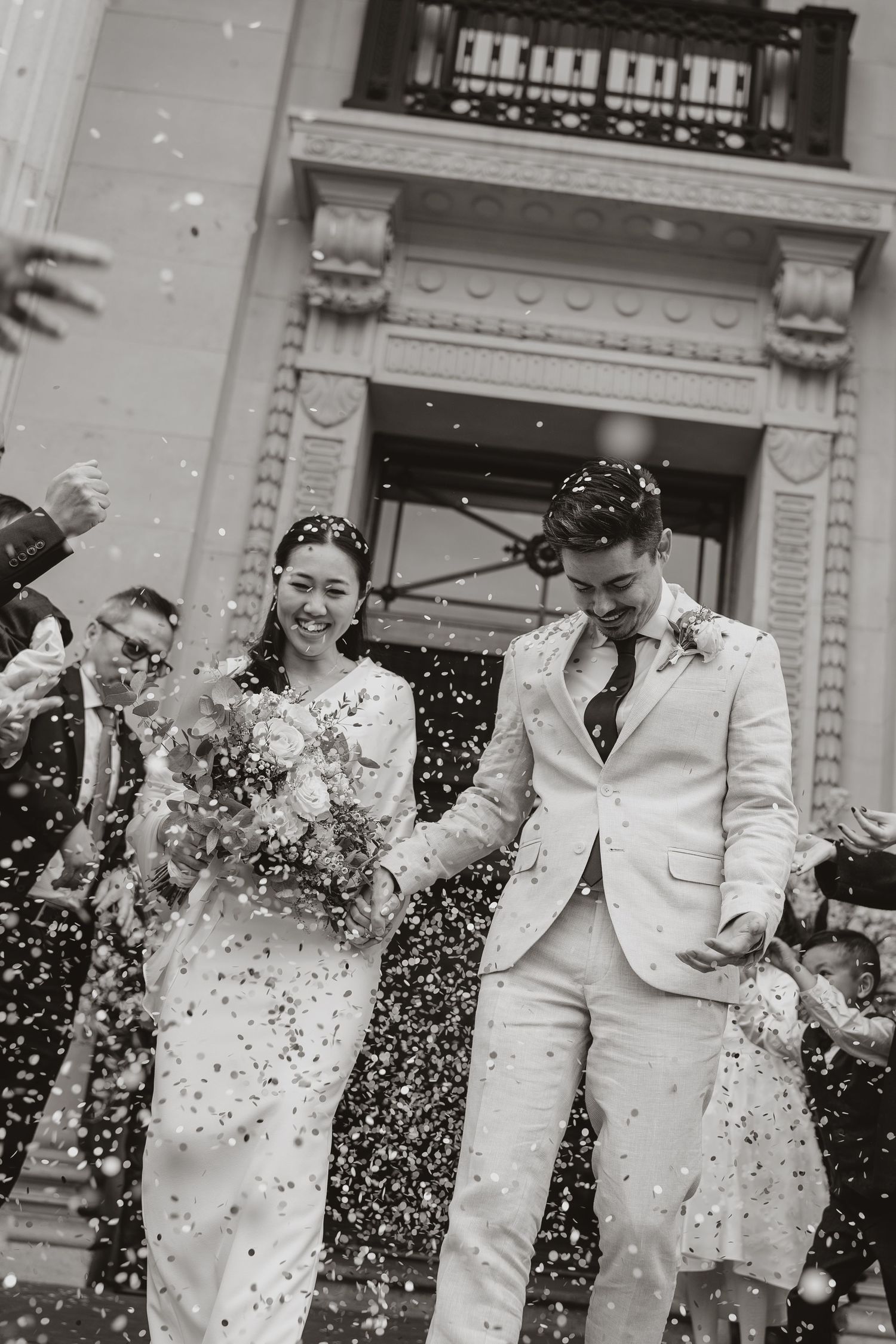 Black and white photo of a couple being showered with confetti as they celebrate their wedding day outside a building.