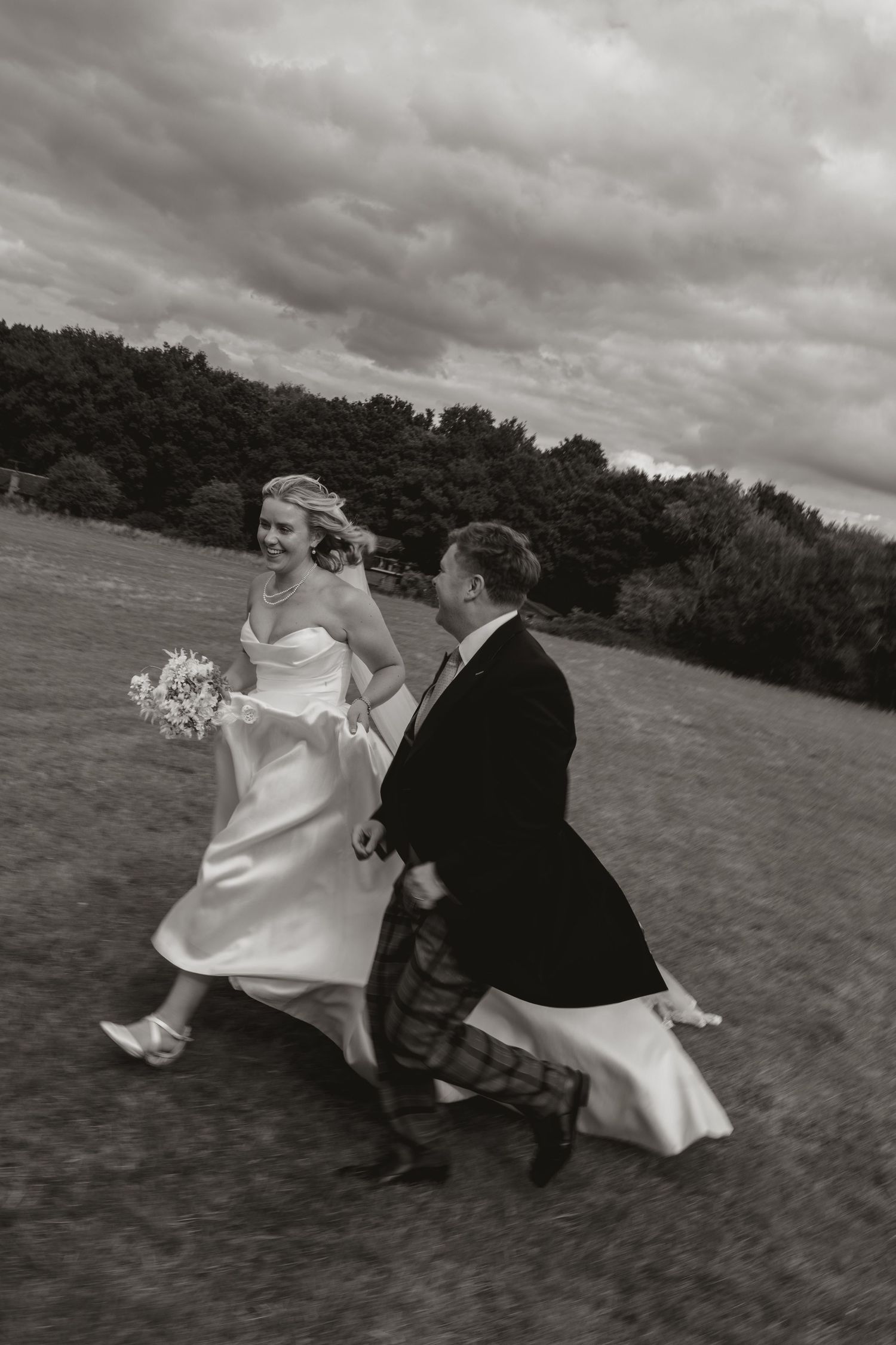 A couple in wedding attire runs playfully across a grassy field under dramatic cloudy skies.
