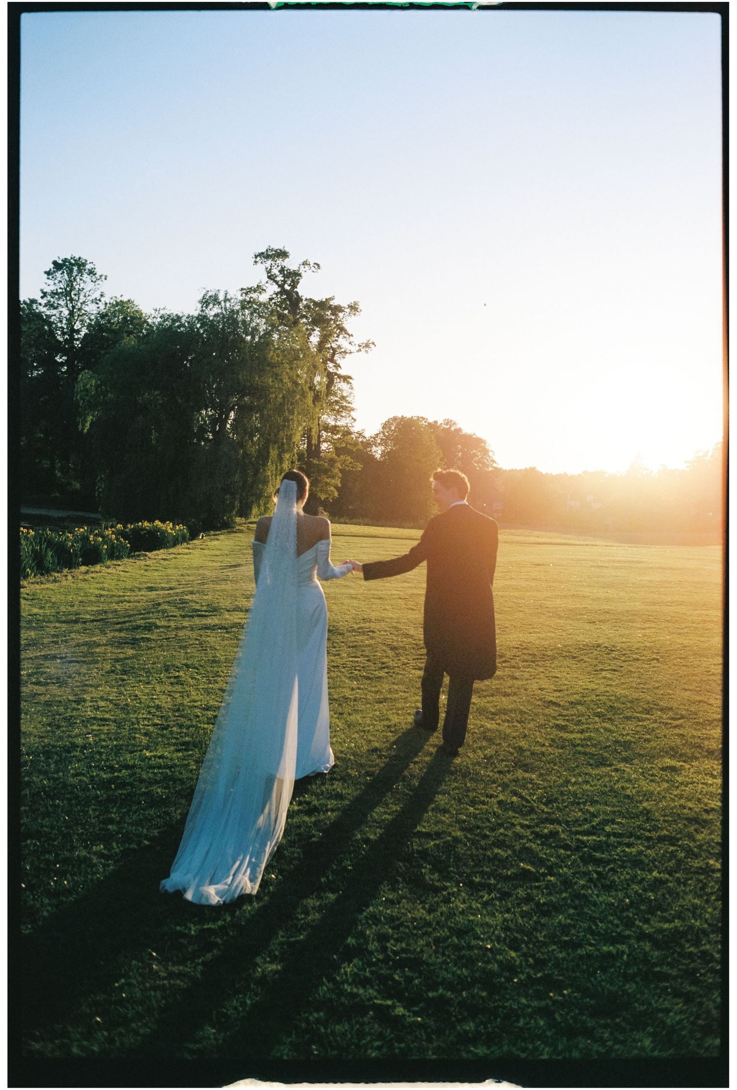 A couple in wedding attire holds hands while walking on a grassy lawn at sunset, casting long shadows.