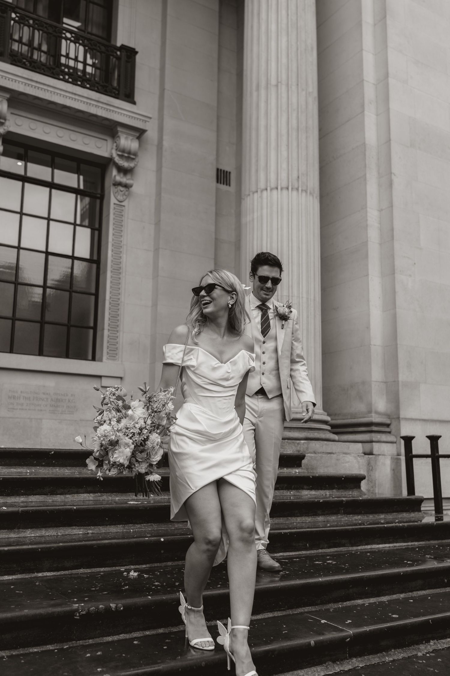 A black and white photo of someone in a white dress running up stone steps of a neoclassical building with columns.