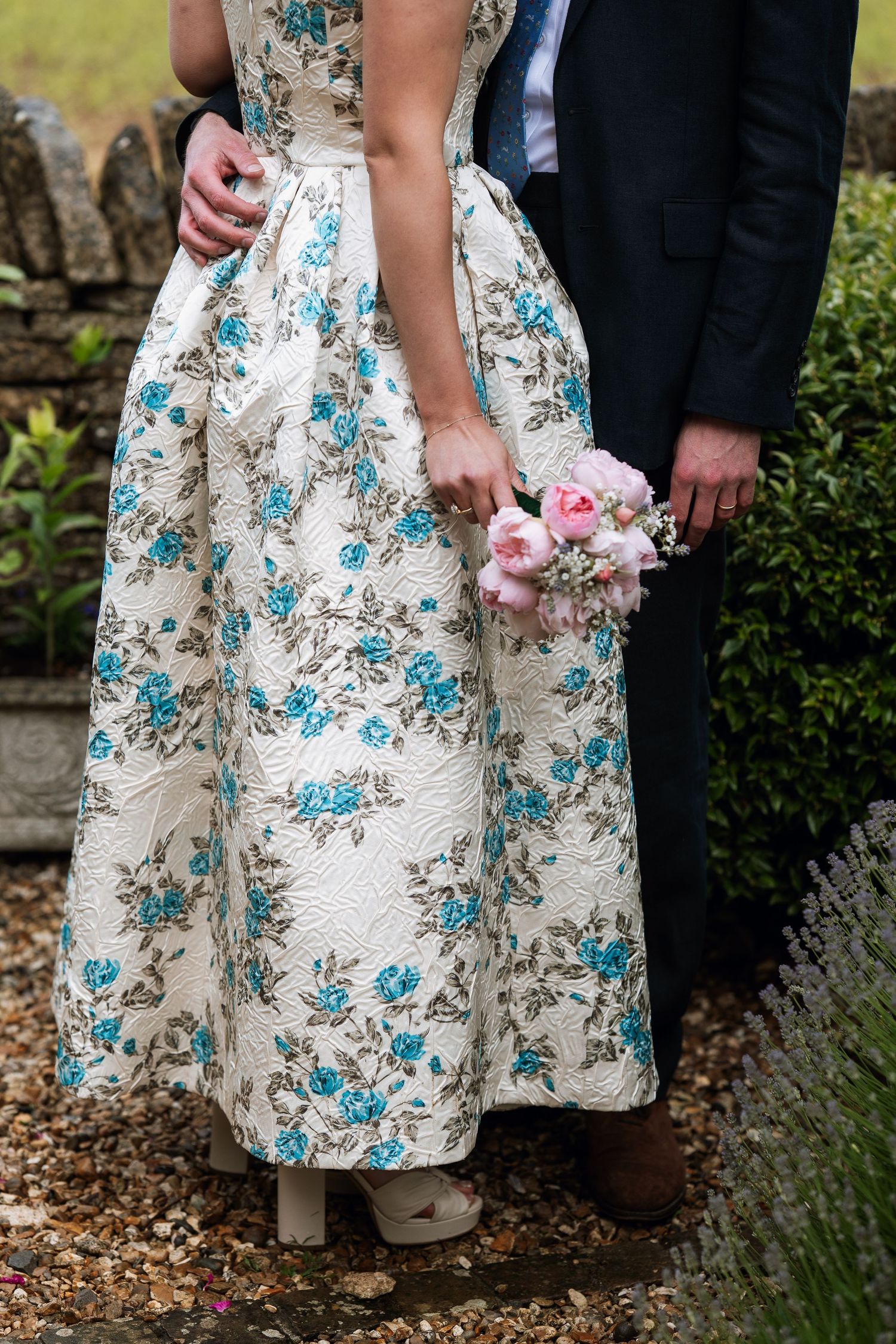 Close-up details show a pattern of turquoise flowers on a white floral dress at an outdoor wedding venue.