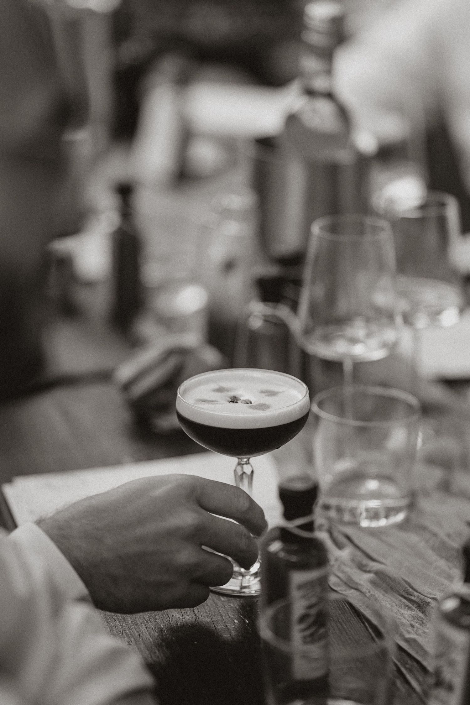A black and white close-up photograph shows hands holding wine glasses and drinks at a social gathering.