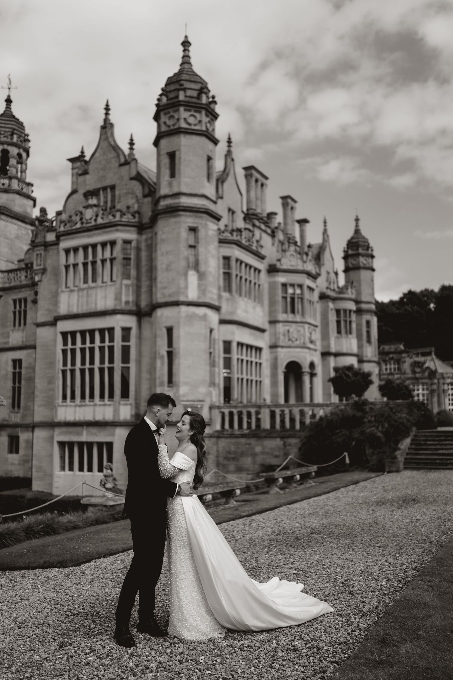 A romantic wedding couple poses in front of a grand castle under dramatic cloudy skies.