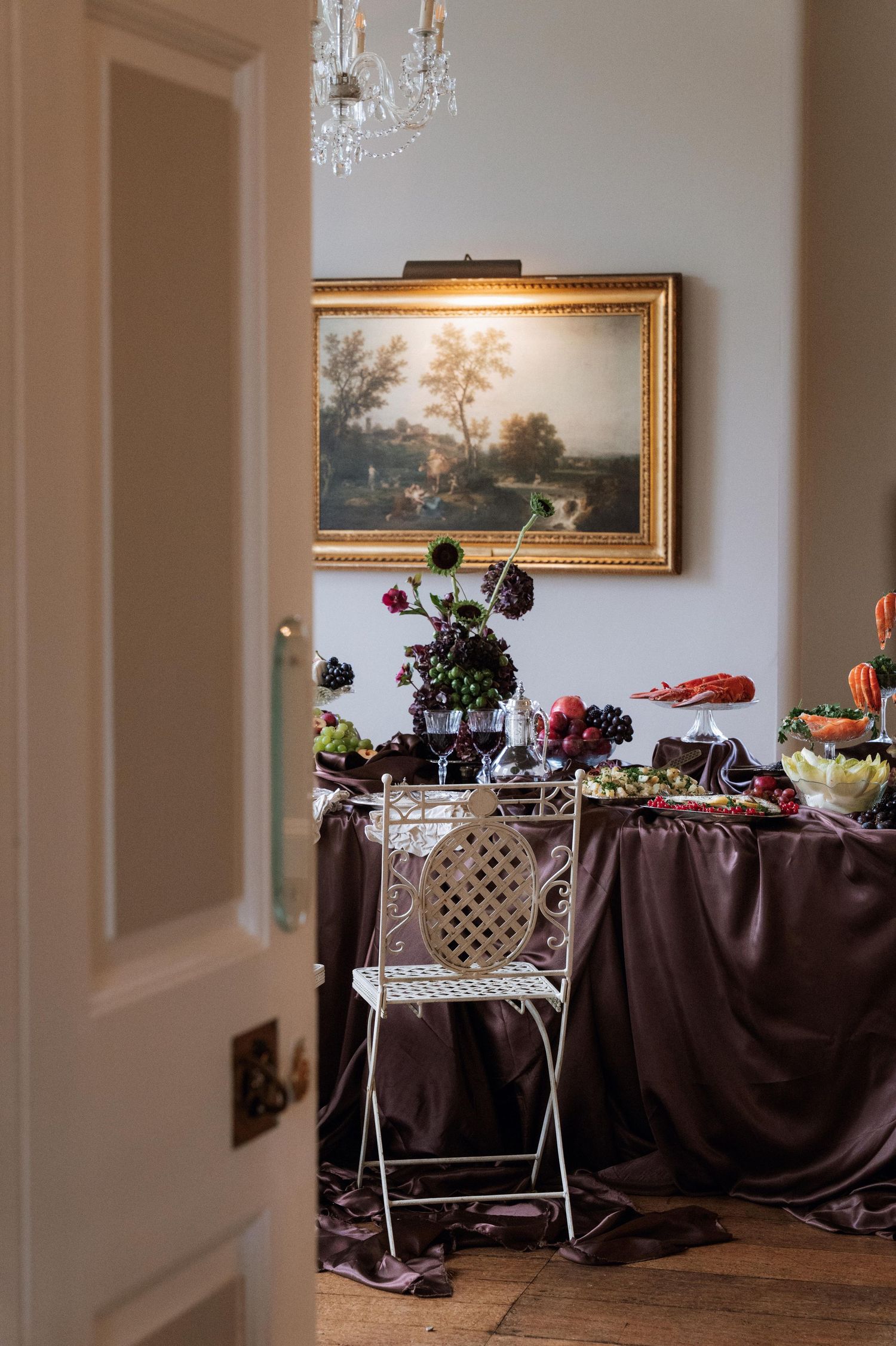 Elegant dining room with framed landscape painting, crystal chandelier, and brown tablecloth set for a formal gathering.