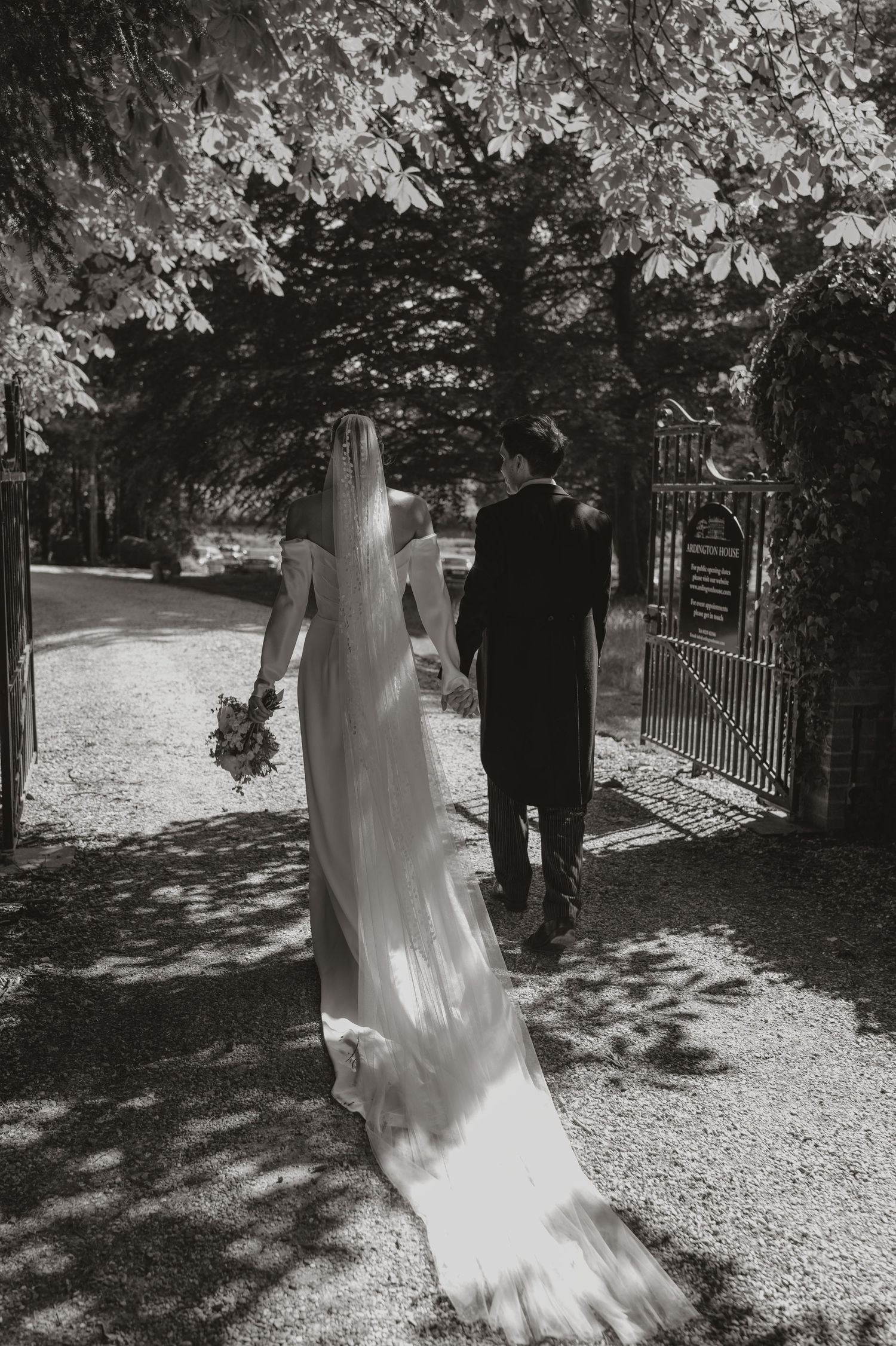 A black and white wedding photo showing a couple walking down a garden path through an ornate gate in dappled sunlight.