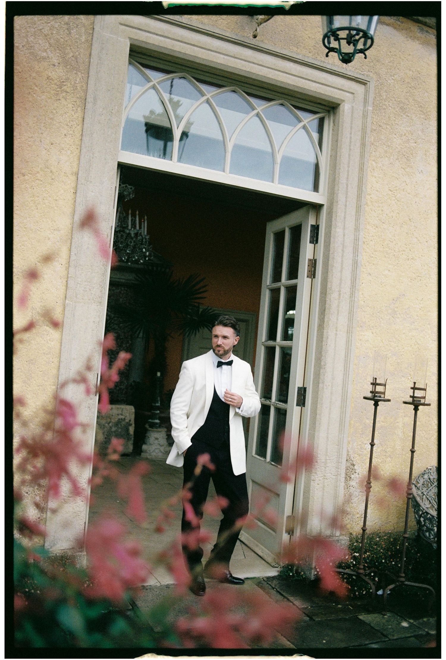 A person in formal attire exits through elegant white double doors of a historic building as pink flowers bloom in foreground.