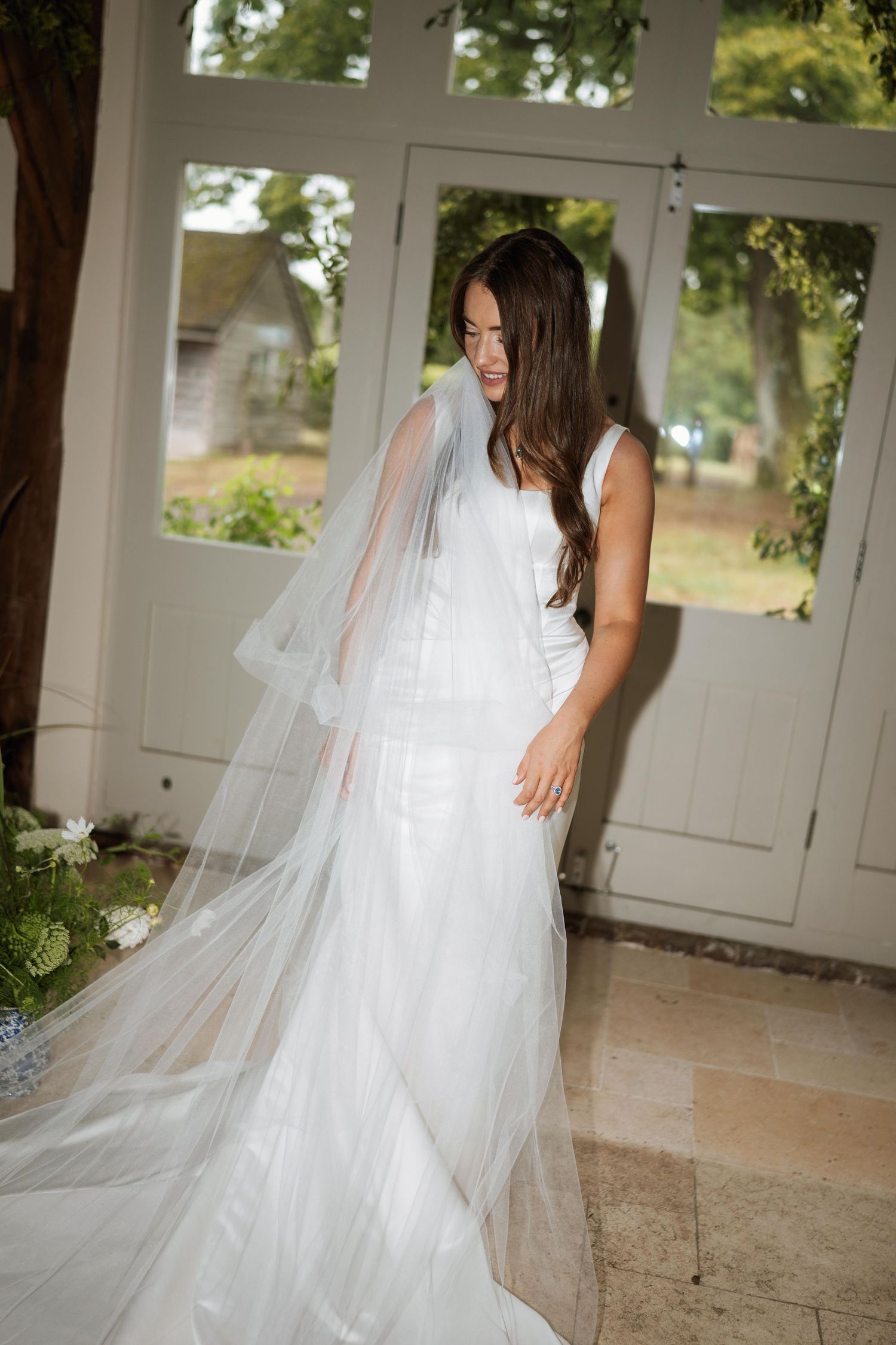 A flowing white wedding gown with delicate tulle veil photographed in a sunlit room with French doors.