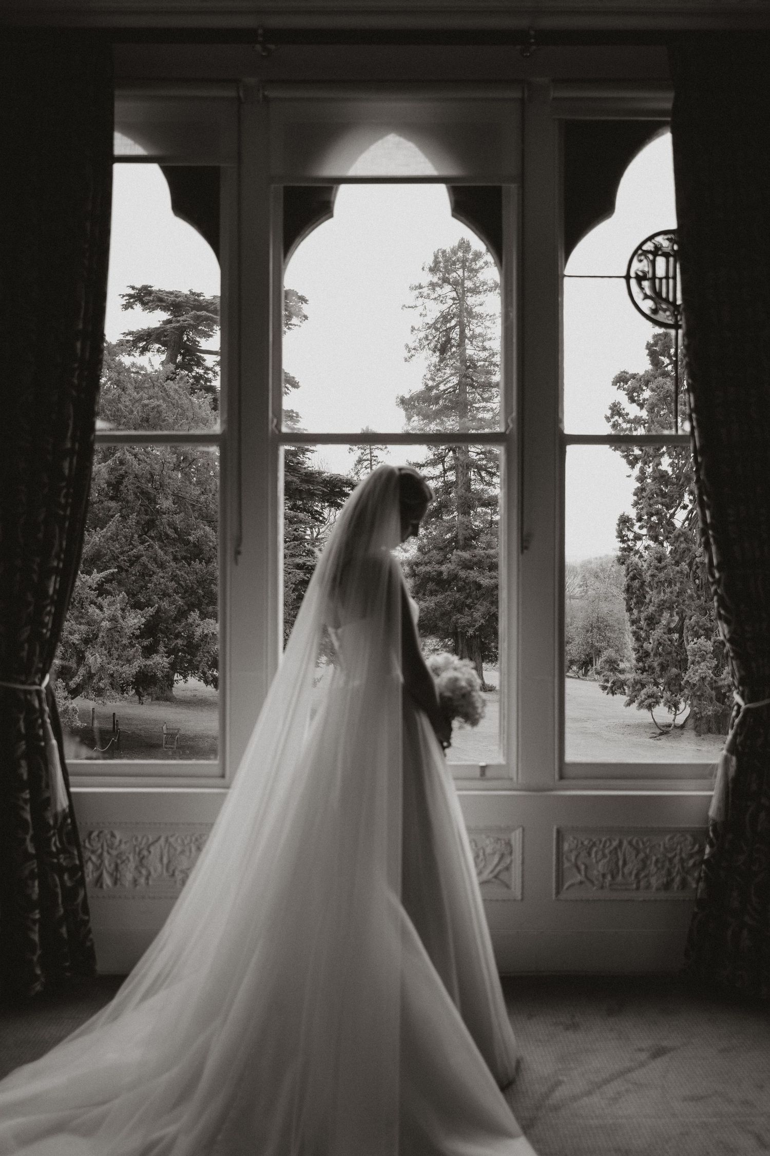 A bride in a flowing white veil stands silhouetted against an arched window overlooking a scenic garden landscape.
