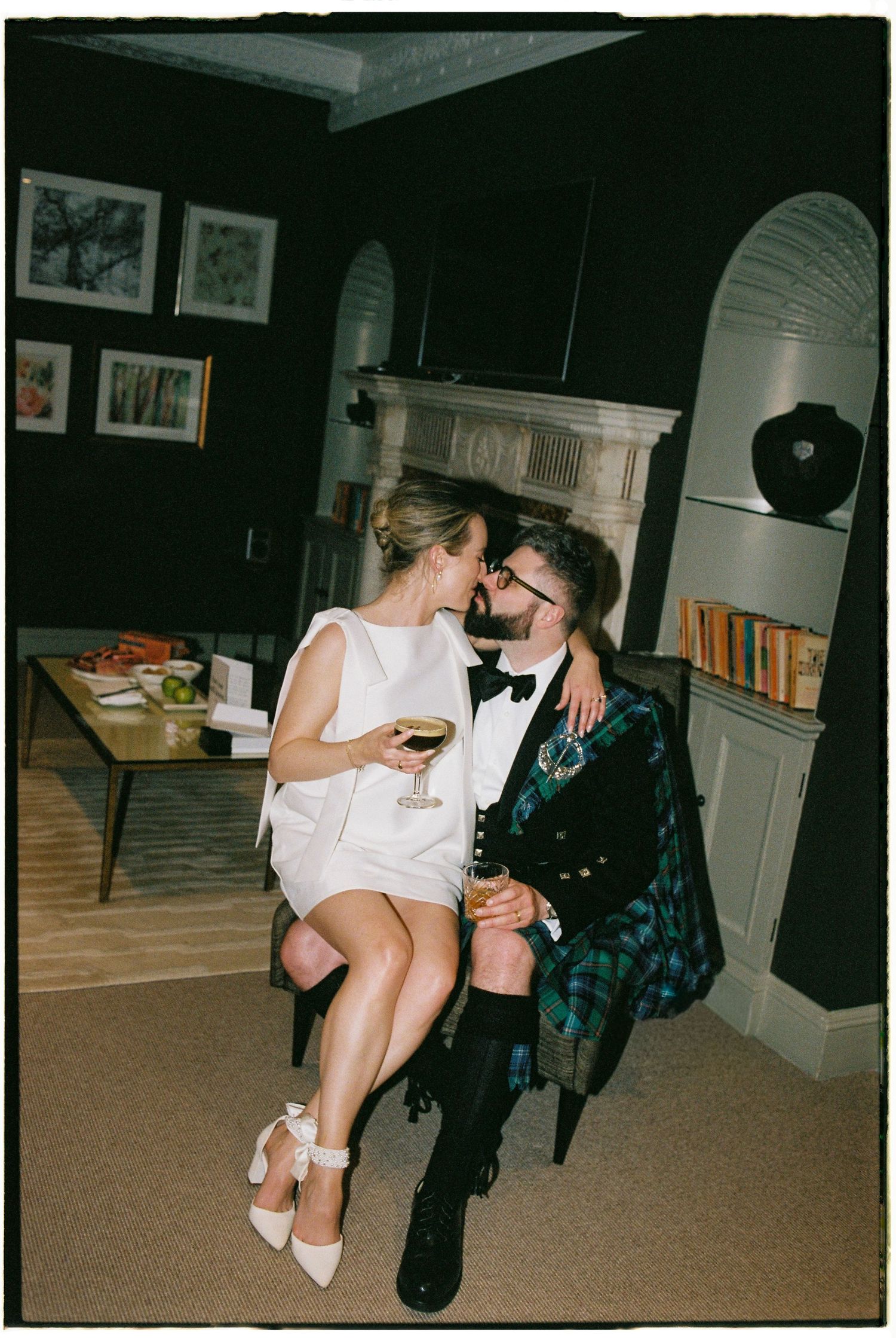 A couple shares an intimate moment at a party, with the woman in white sitting on a table and holding a drink.