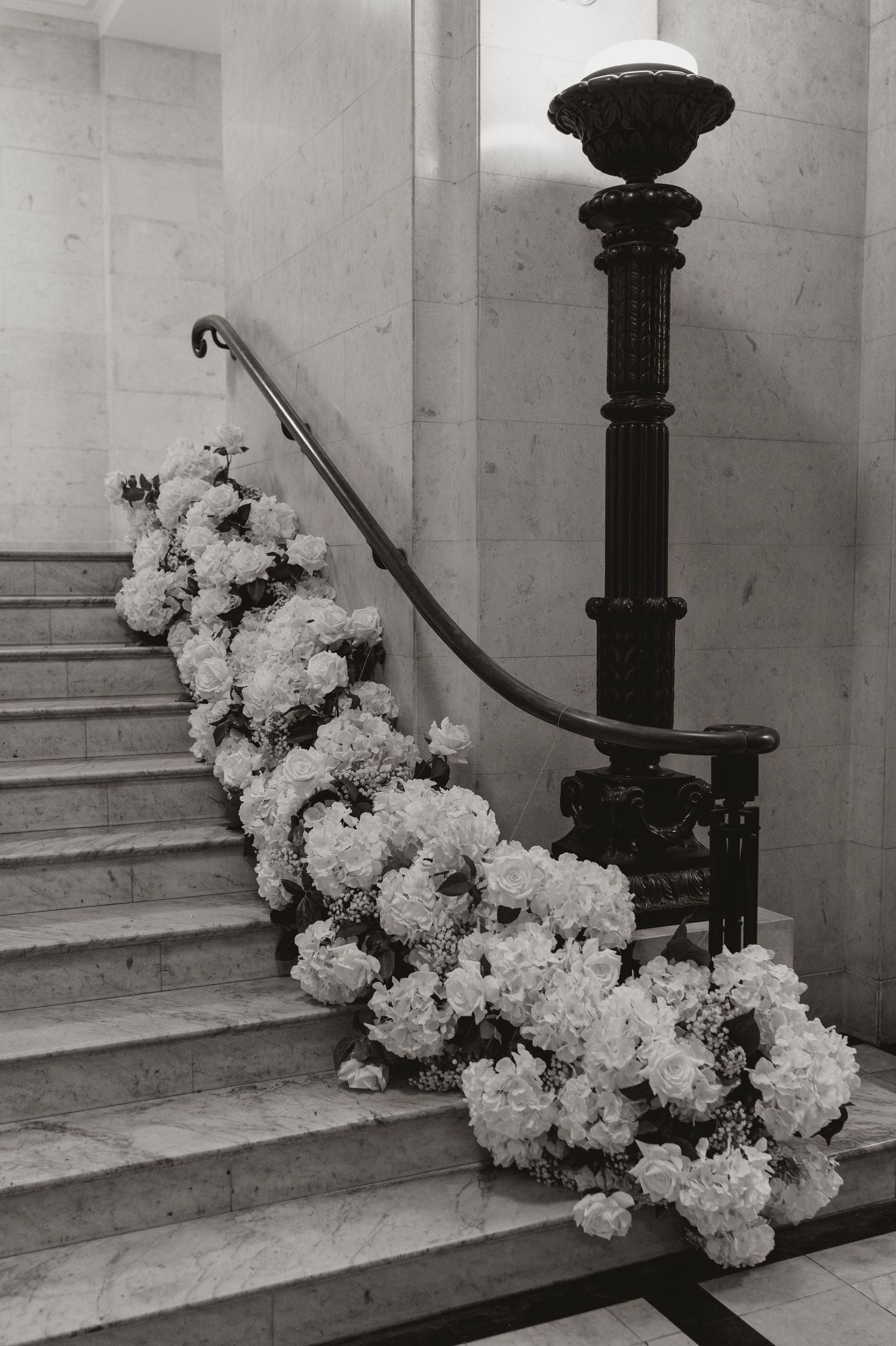 Black and white photograph of white hydrangea flowers cascading down marble stairs with ornate iron railings.