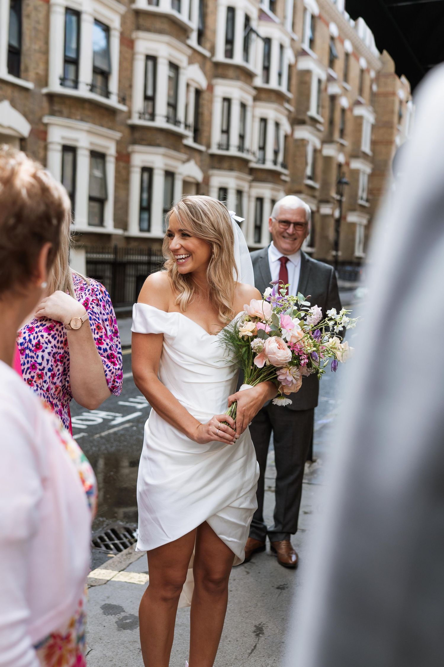 Bride in white dress and guest in pink floral dress share joyful moments on city street.